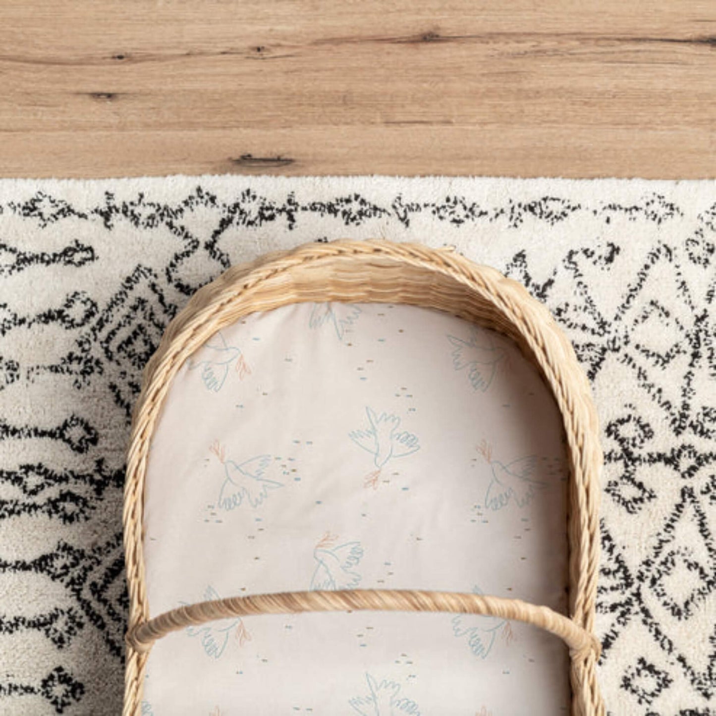 A Moses basket lined with a cream fitted sheet featuring a subtle bird print, placed on a patterned rug and wooden floor.
