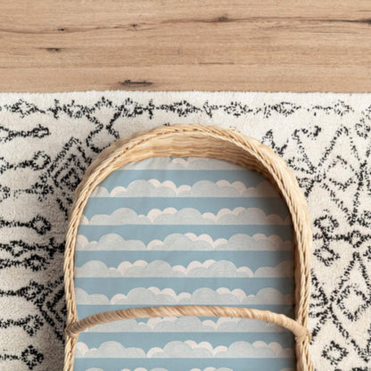 Moses basket lined with a blue rolling cloud fitted sheet, shown inside a woven palm leaf basket on a patterned rug.