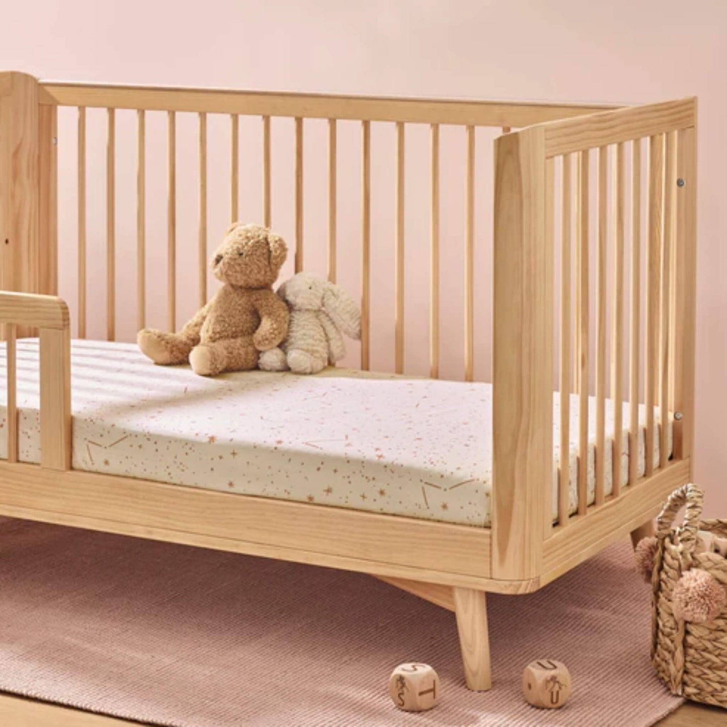 Wooden cot styled with a cream fitted sheet featuring scattered stars and constellation lines, with two soft toys placed on the mattress.