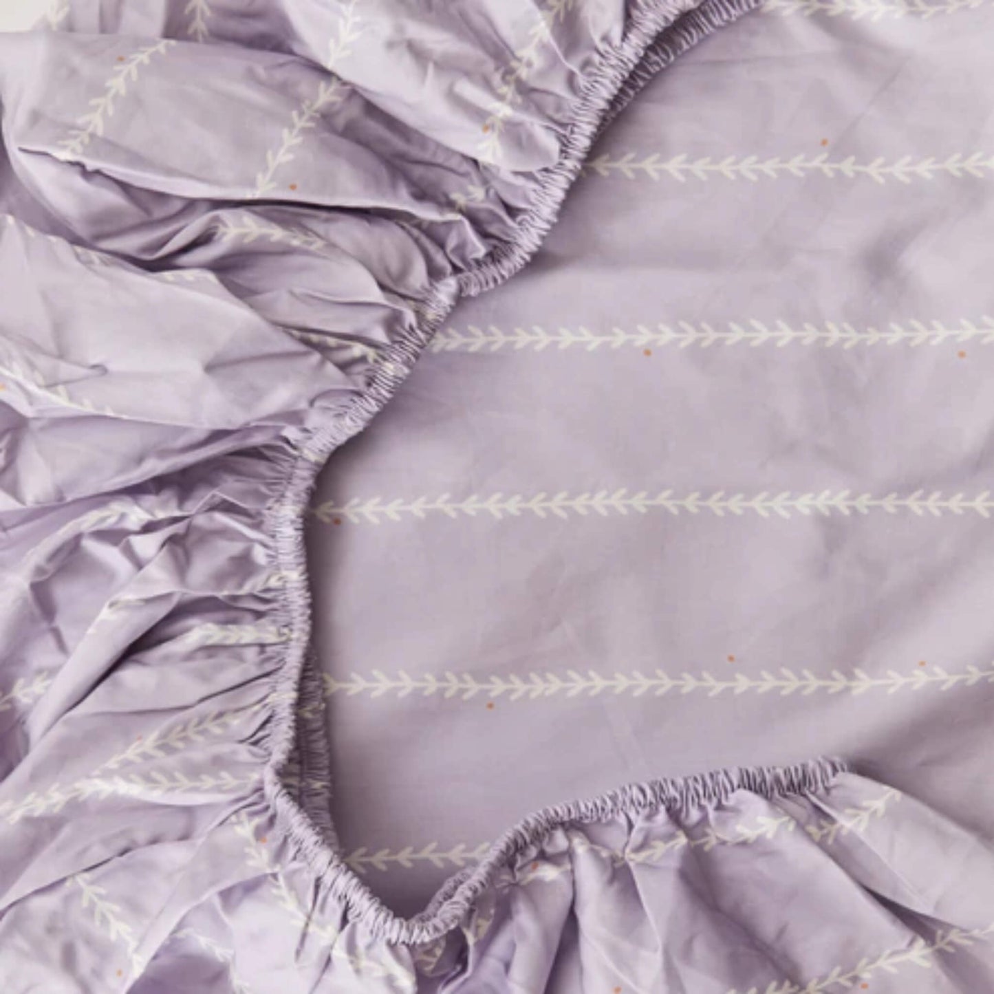 A close-up of elasticated edges on a lilac fitted sheet, highlighting gathered fabric and stitched vine detailing.
