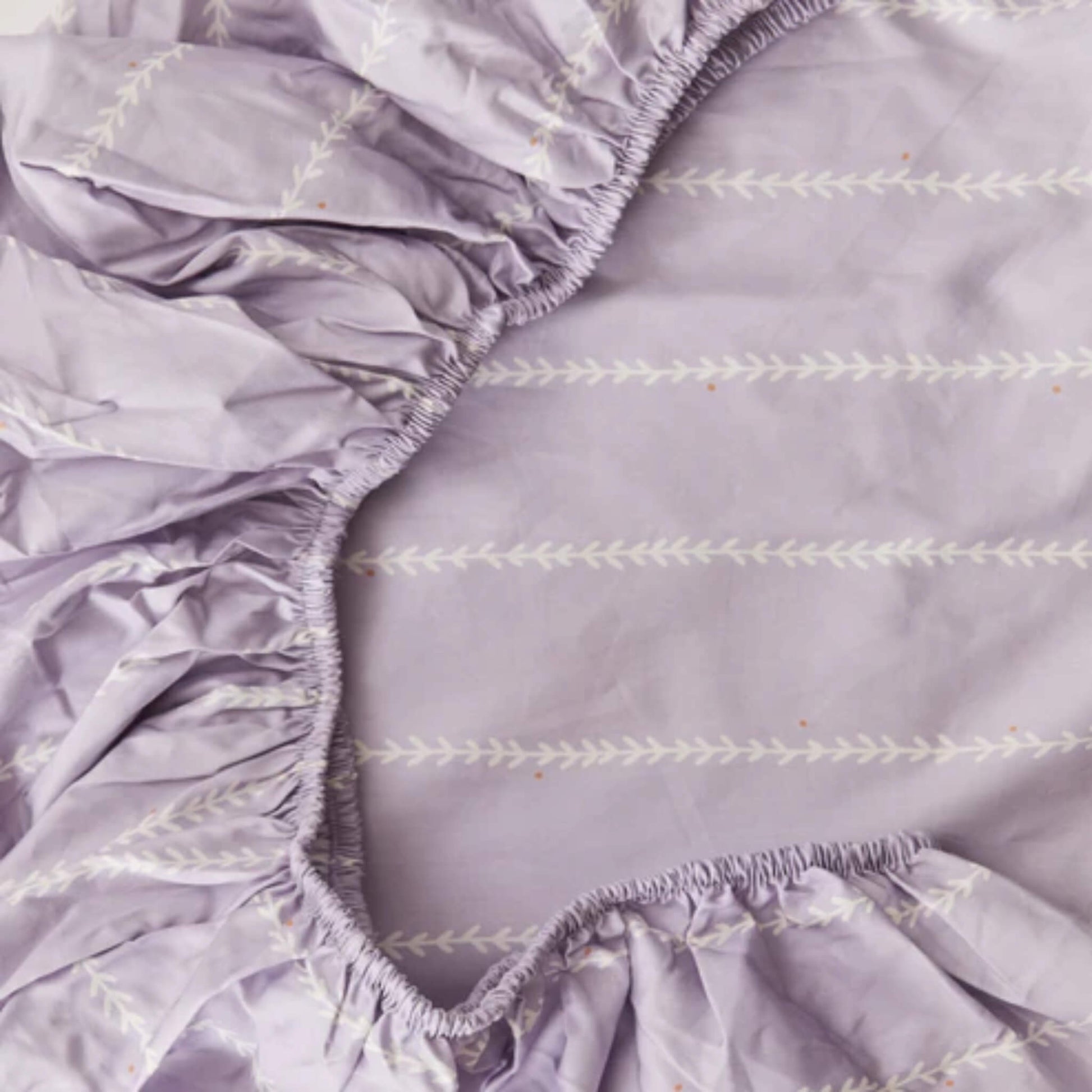 A close-up of elasticated edges on a lilac fitted sheet, highlighting gathered fabric and stitched vine detailing.