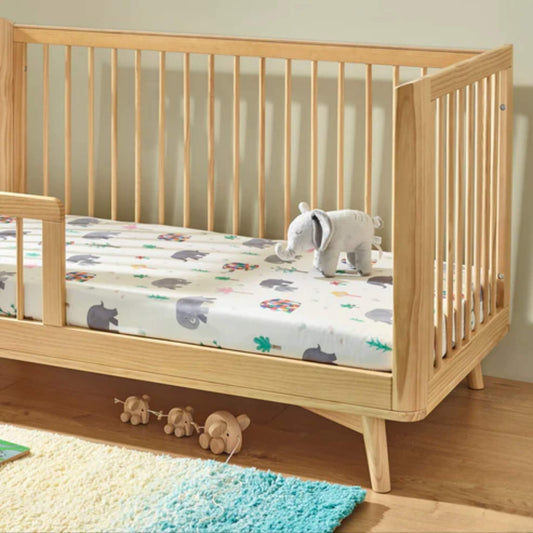 A cot bed mattress dressed with a fitted sheet printed with grey elephants, colourful patchwork elephants, and small trees, photographed inside a wooden cot bed.