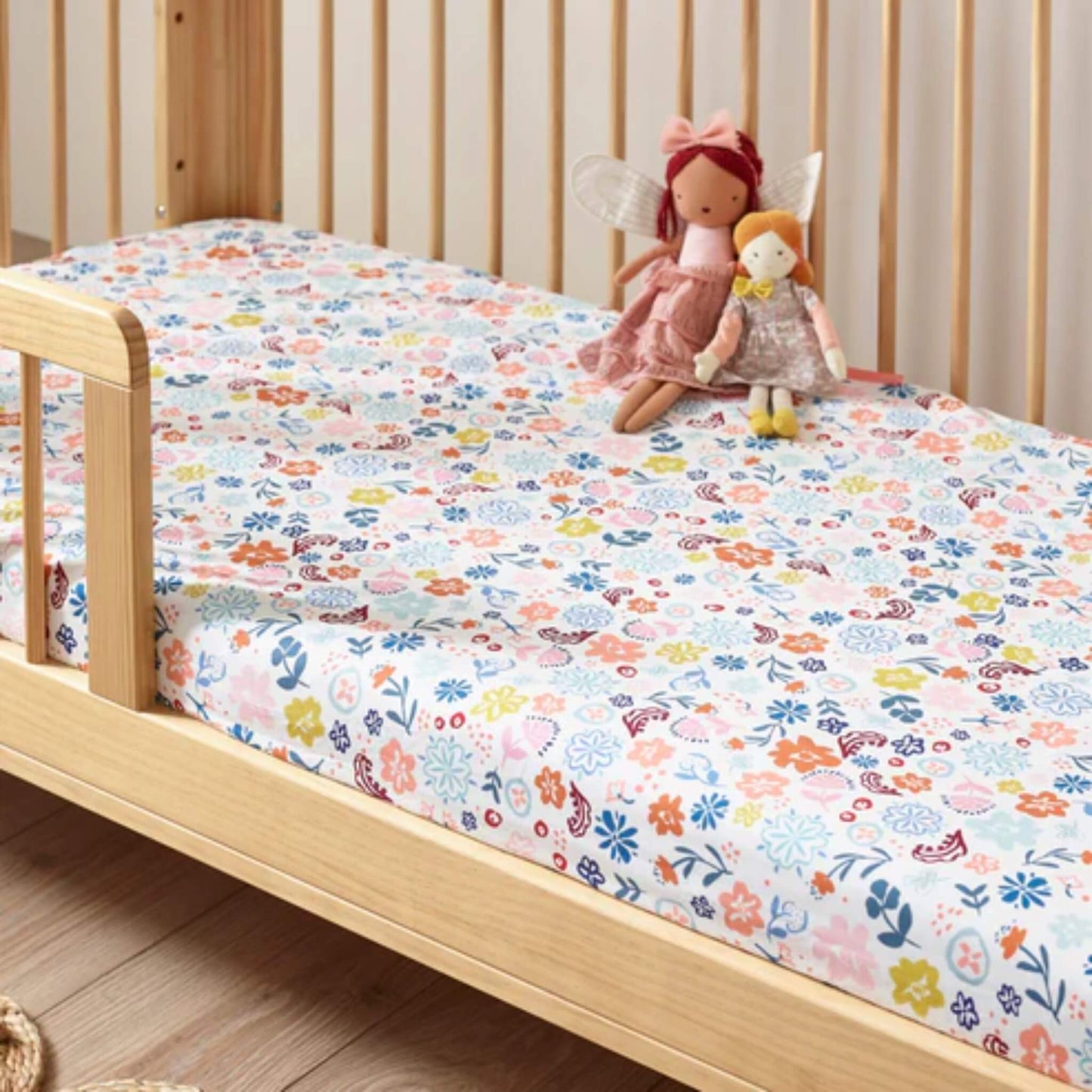 A fitted cotton sheet on a wooden cot bed, featuring an all-over floral pattern in soft pastel and brighter accent colours.