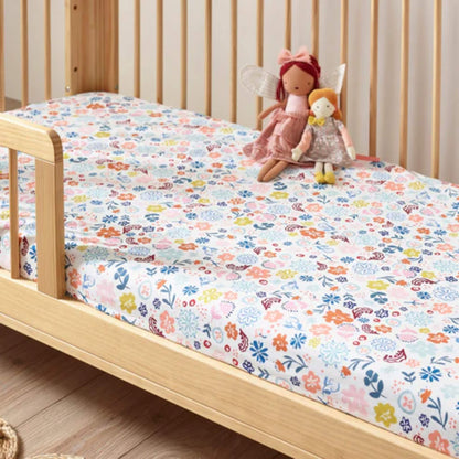 A fitted cotton sheet on a wooden cot bed, featuring an all-over floral pattern in soft pastel and brighter accent colours.