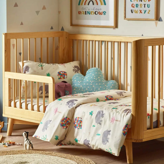 A cot bed dressed with a cream duvet cover and pillowcase featuring Elmer patchwork elephants, styled in a warm wooden nursery with soft wall art and toys.