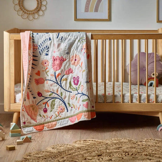 A quilt draped over a wooden cot rail, showing a detailed floral and animal print with soft pink, green, and blue tones in a calm nursery setting.