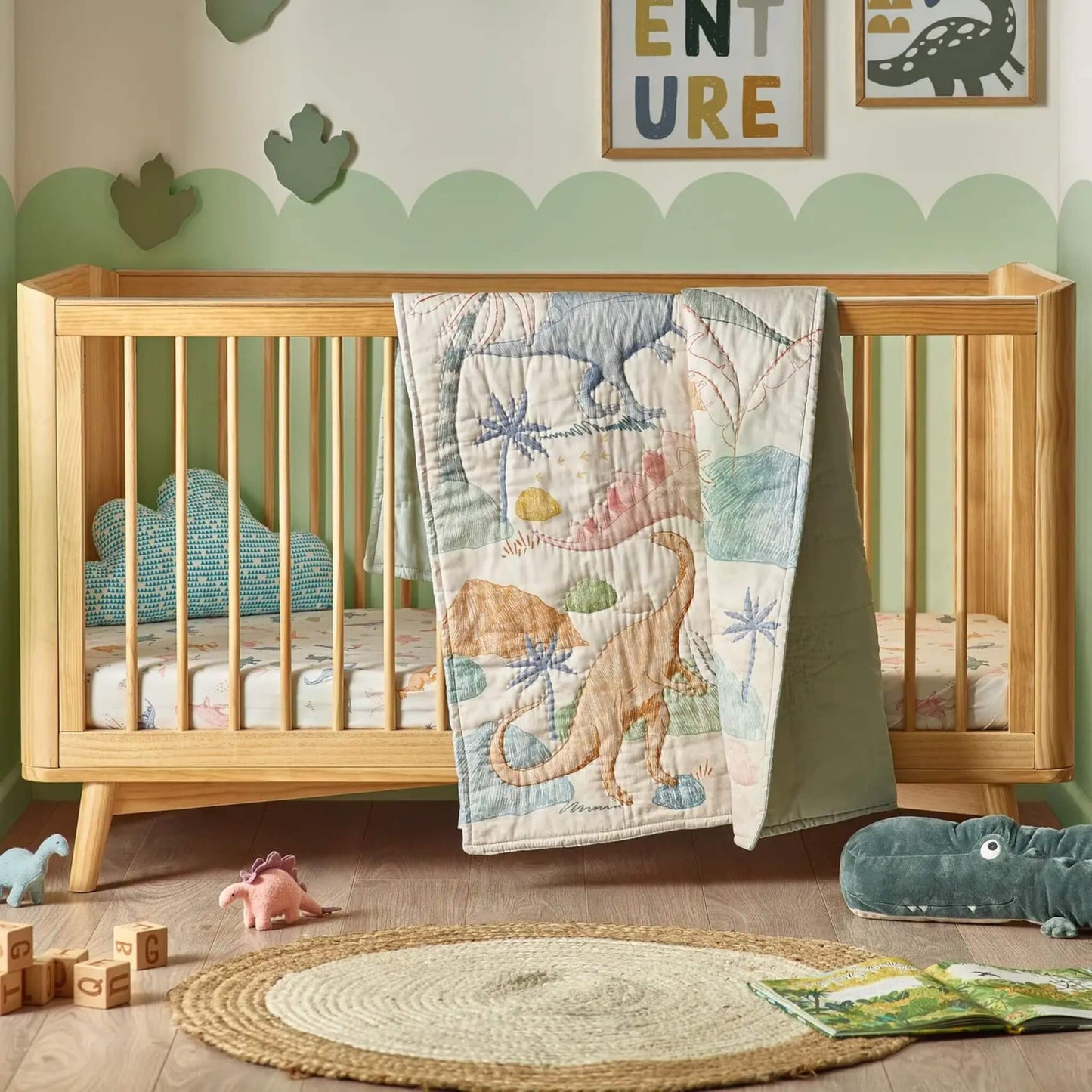 A wooden cot bed with a dinosaur-patterned quilt draped over the rail, showing large illustrated dinosaurs in muted greens, blues, and warm neutrals.