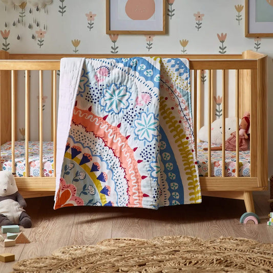 A wooden cot bed with a floral quilt draped over a side rail, showing curved patterned bands in pastel blue, coral, mustard, and soft white.