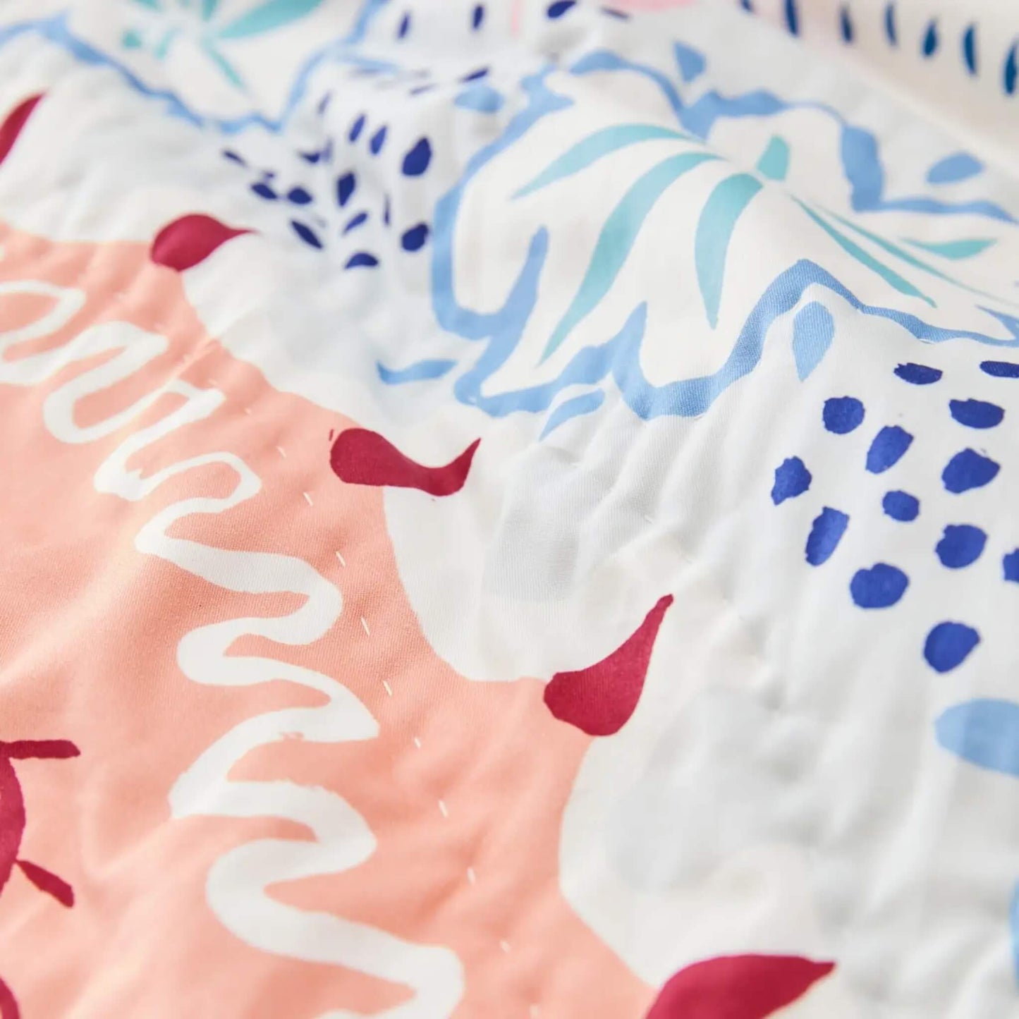 A detailed view of the quilt surface showing stitched cotton texture, curved decorative lines, and floral motifs in coral, blue, and soft red accents.