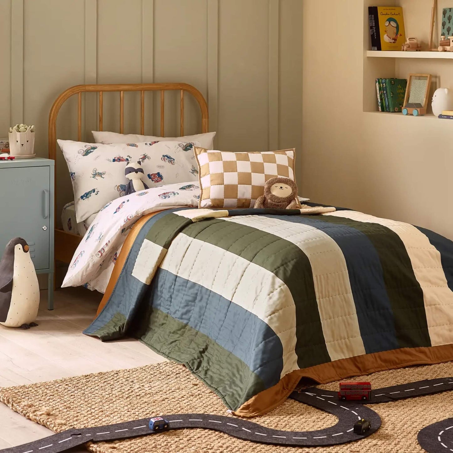 A child’s single bed styled with a striped quilt layered over vehicle-themed bedding, featuring broad vertical stripes in muted blue, green, and cream tones.
