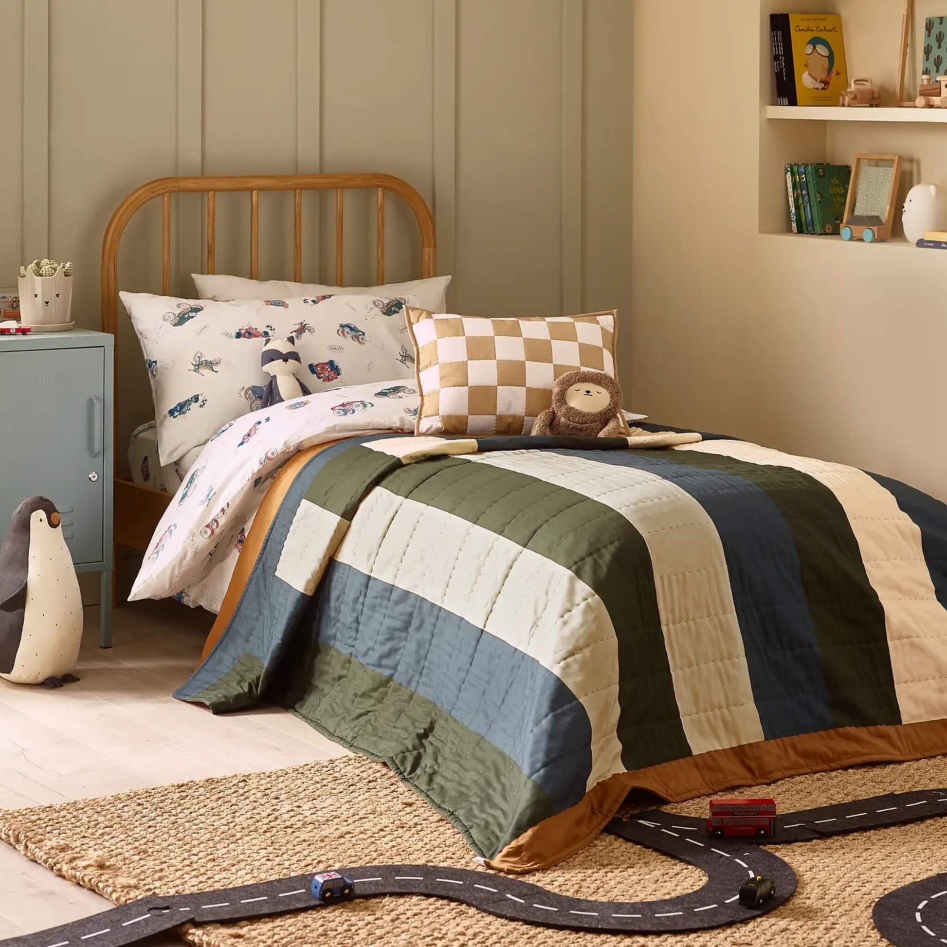 A child’s single bed styled with a striped quilt layered over vehicle-themed bedding, featuring broad vertical stripes in muted blue, green, and cream tones.