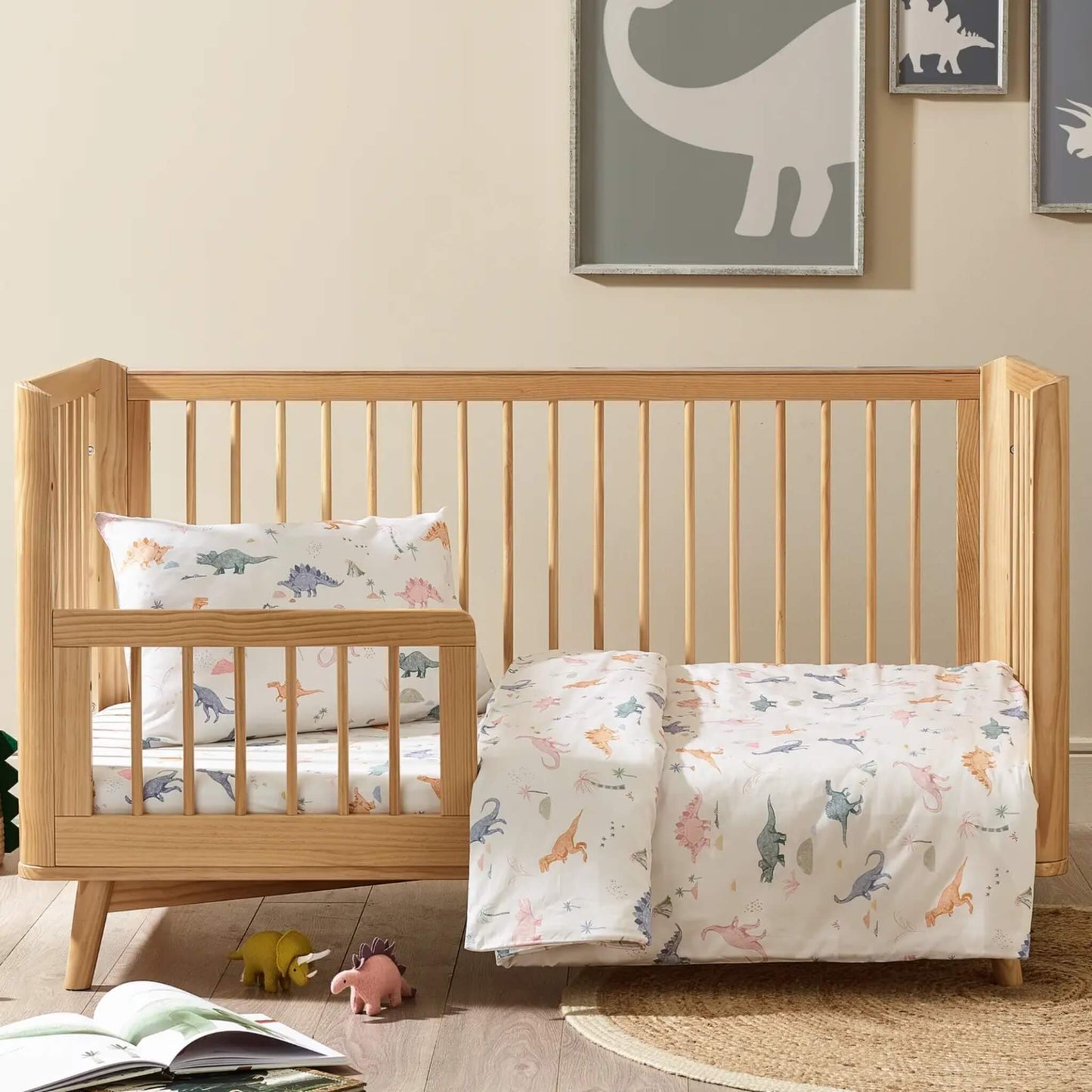 Dino print Kabode cot bed duvet cover and pillowcase set in a soft cream organic cotton printed with hand-drawn dinosaurs in muted pastel tones, shown folded neatly with the matching pillowcase on top.