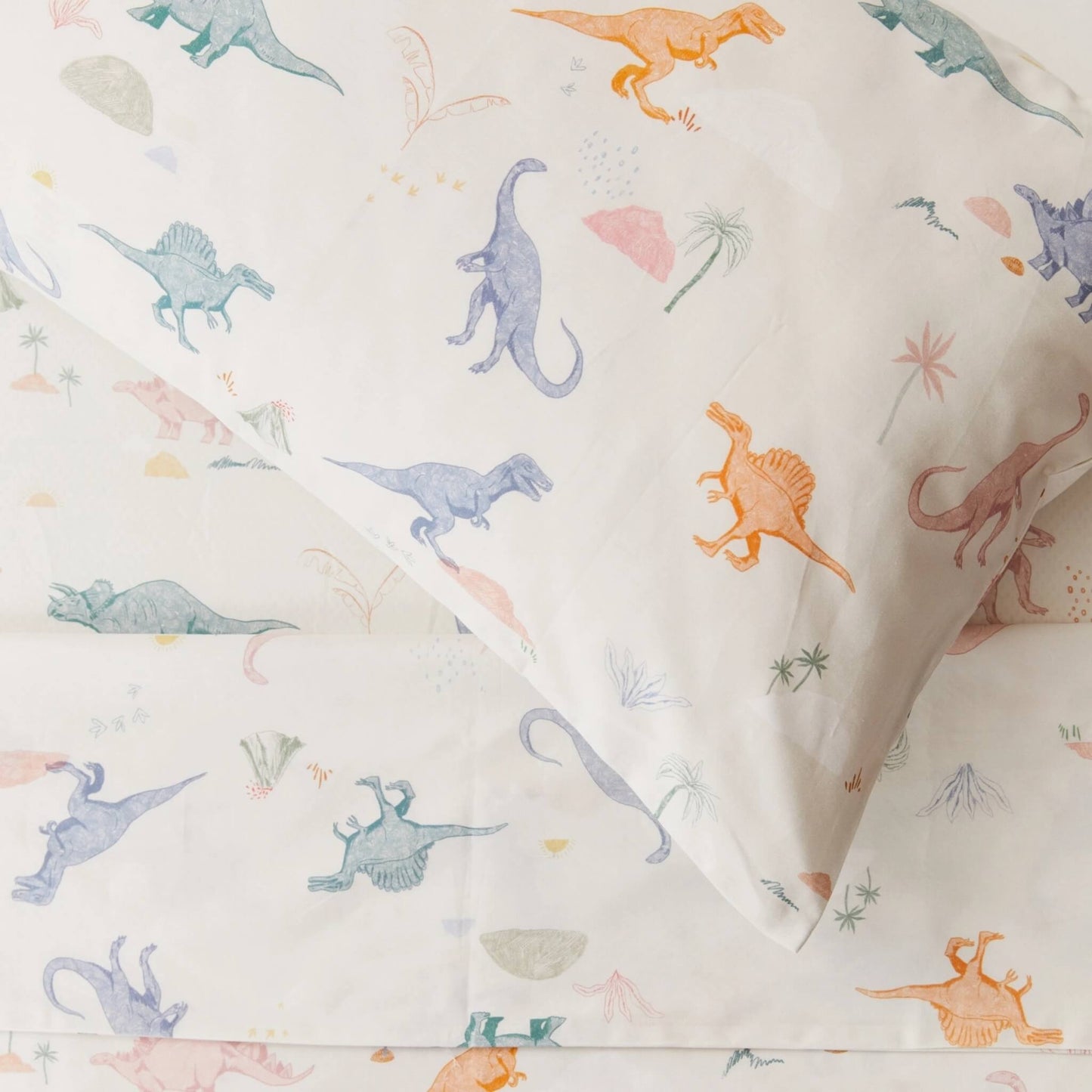Close-up view of a pillowcase printed with illustrated dinosaurs in muted blues, greens, and warm tones on light cream cotton.