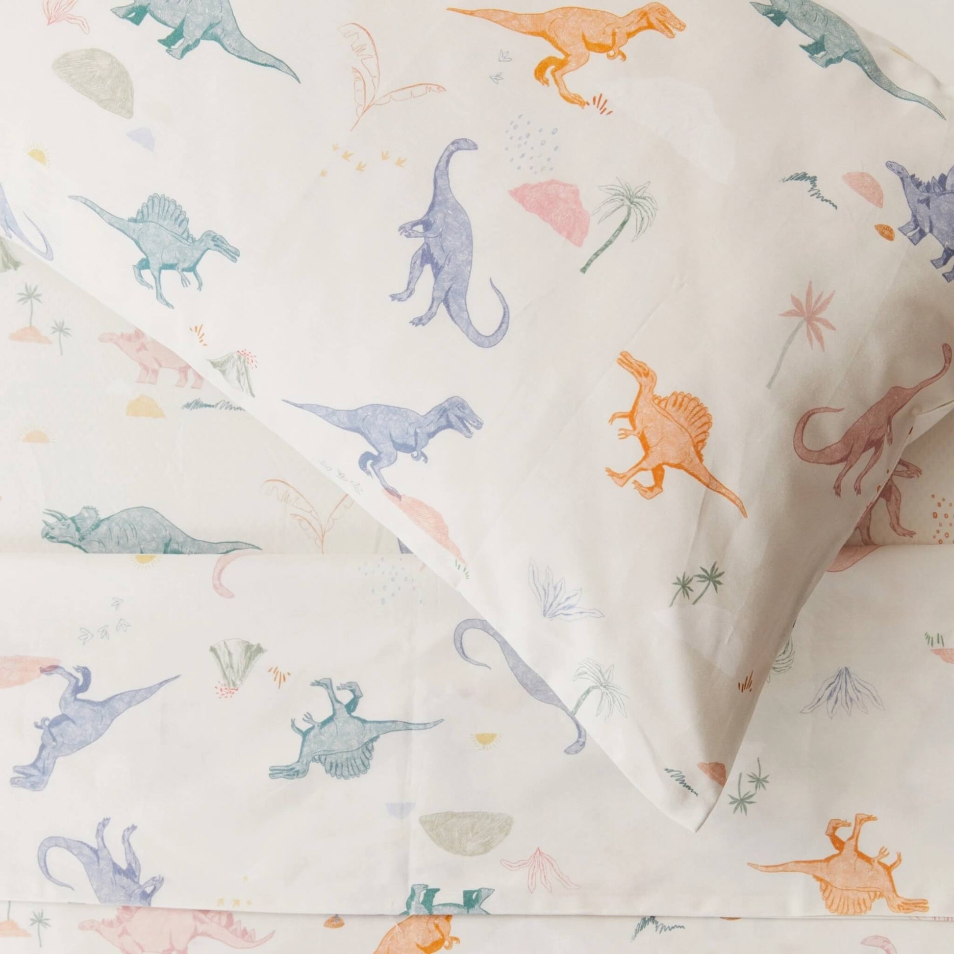 Close-up view of a pillowcase printed with illustrated dinosaurs in muted blues, greens, and warm tones on light cream cotton.