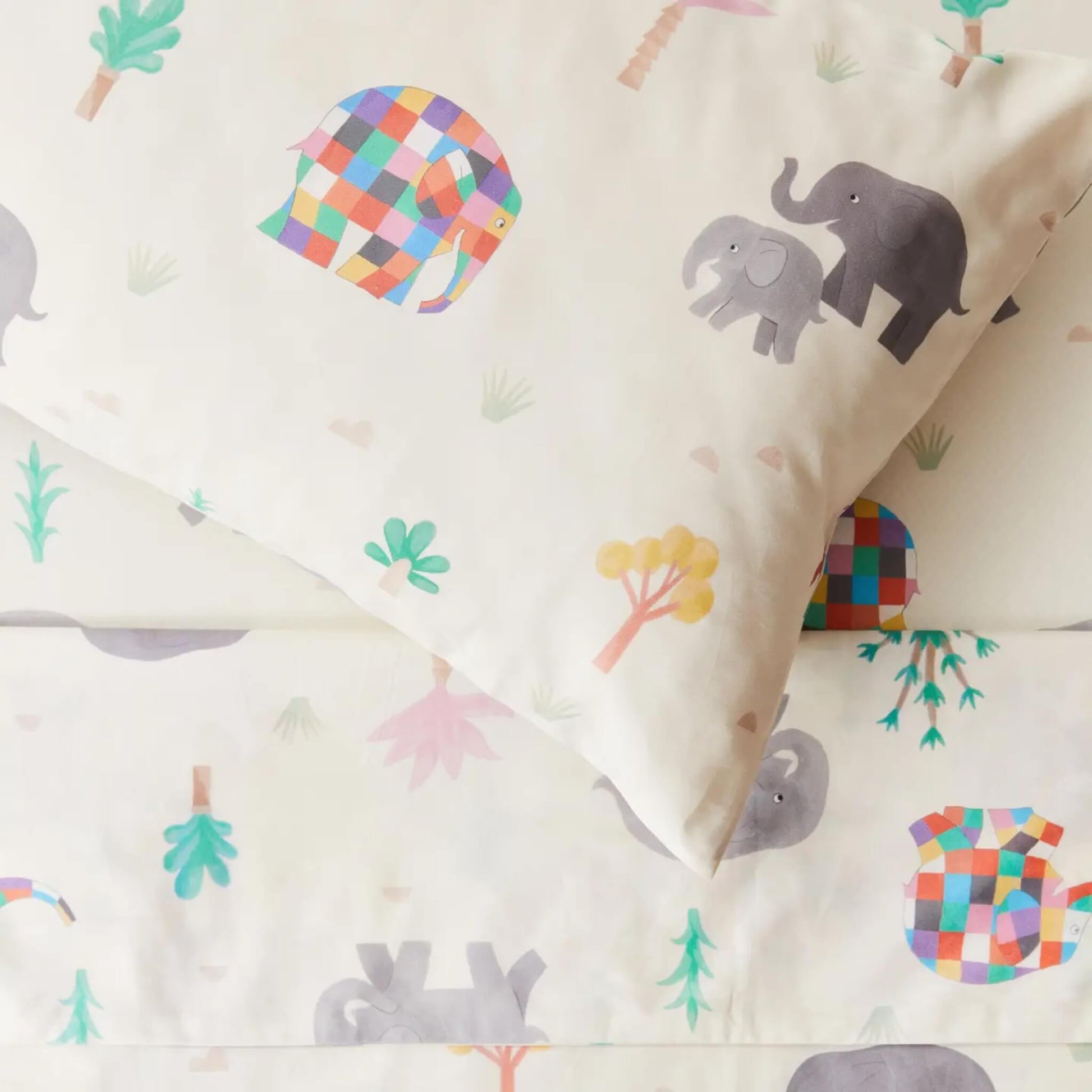 A close-up of a pillowcase and duvet cover showing Elmer patchwork elephants, small trees, and soft pastel details printed on smooth cream cotton fabric.