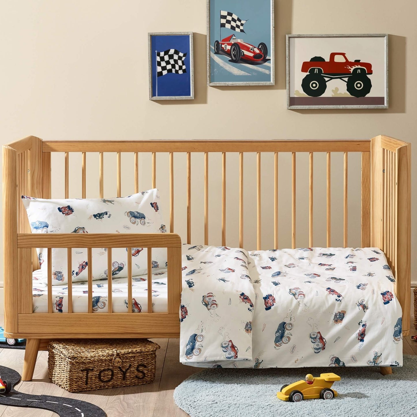 Kabode cot bed duvet cover and pillowcase set in a vehicles print, shown on a wooden cot bed with a white base fabric illustrated with cars, motorbikes, trucks, flags, and playful motion details.