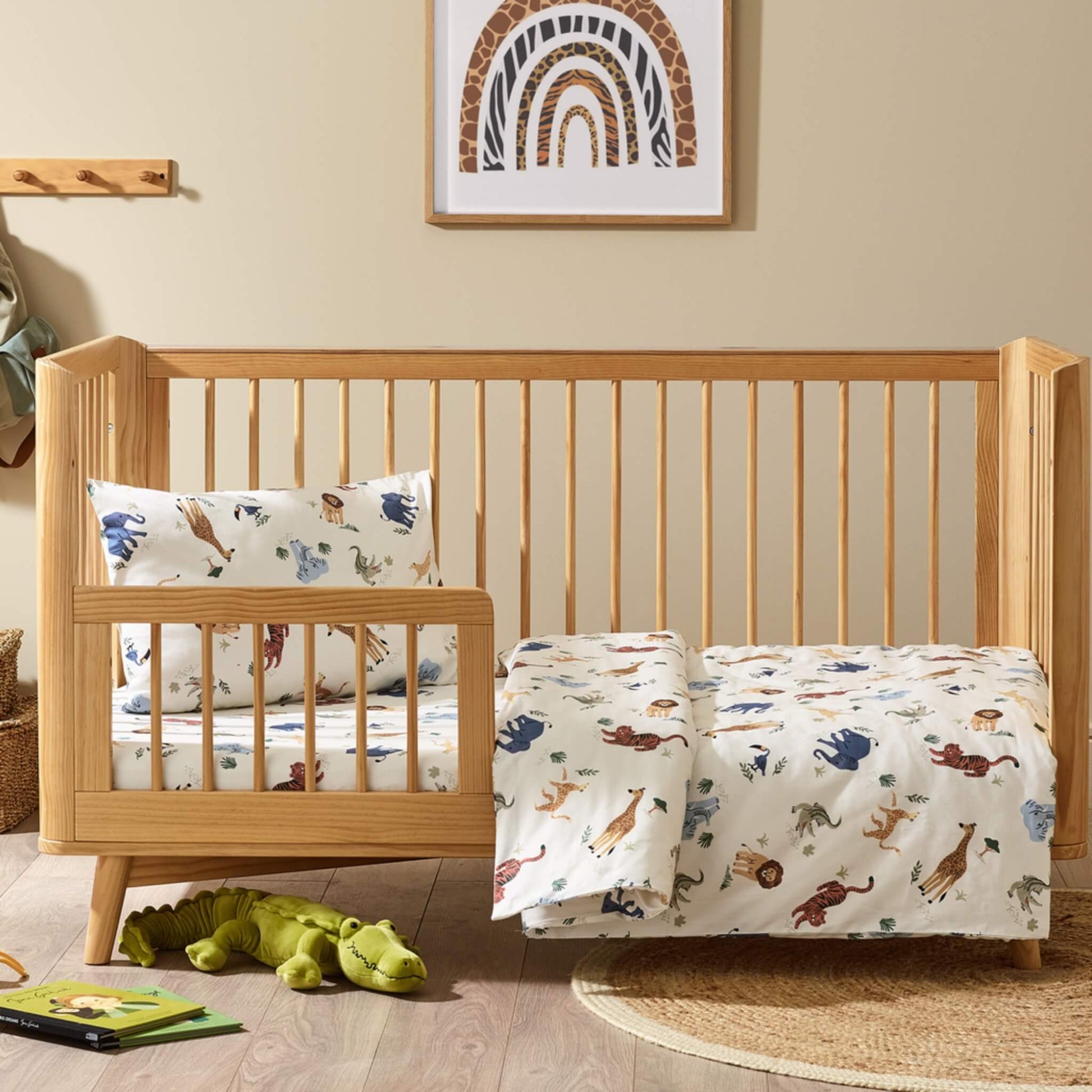 Kabode safari print cot bed duvet cover and pillowcase set styled on a wooden cot bed, with cream bedding decorated with illustrated jungle animals including lions, elephants, giraffes and tigers in a calm nursery setting.