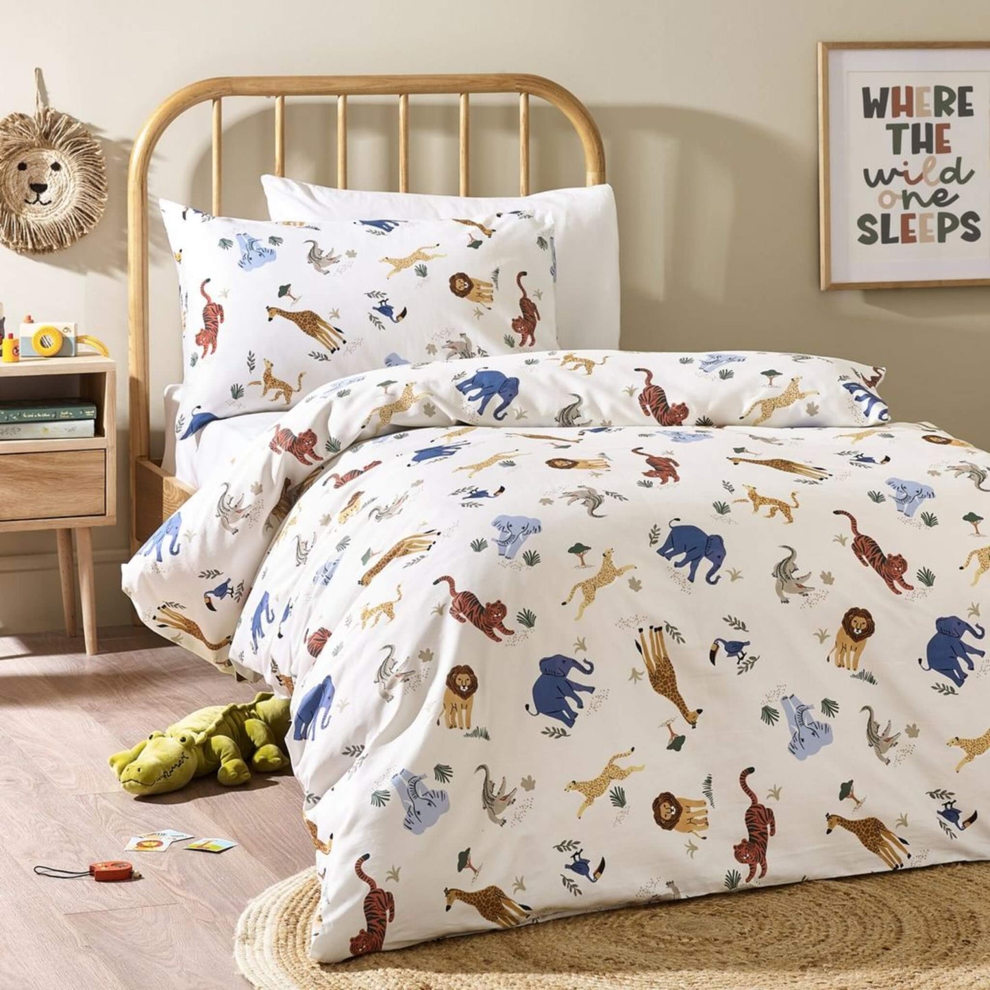 A child’s single bed dressed in cream safari-themed bedding featuring illustrated jungle animals such as elephants, lions, giraffes and tigers, styled with a wooden bed frame and soft nursery accessories.