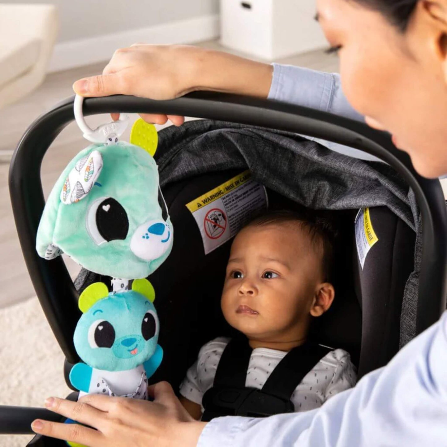 Baby seated in a car seat gazing at a hanging plush bear toy clipped to the handle above, engaging in visual and sensory play.
