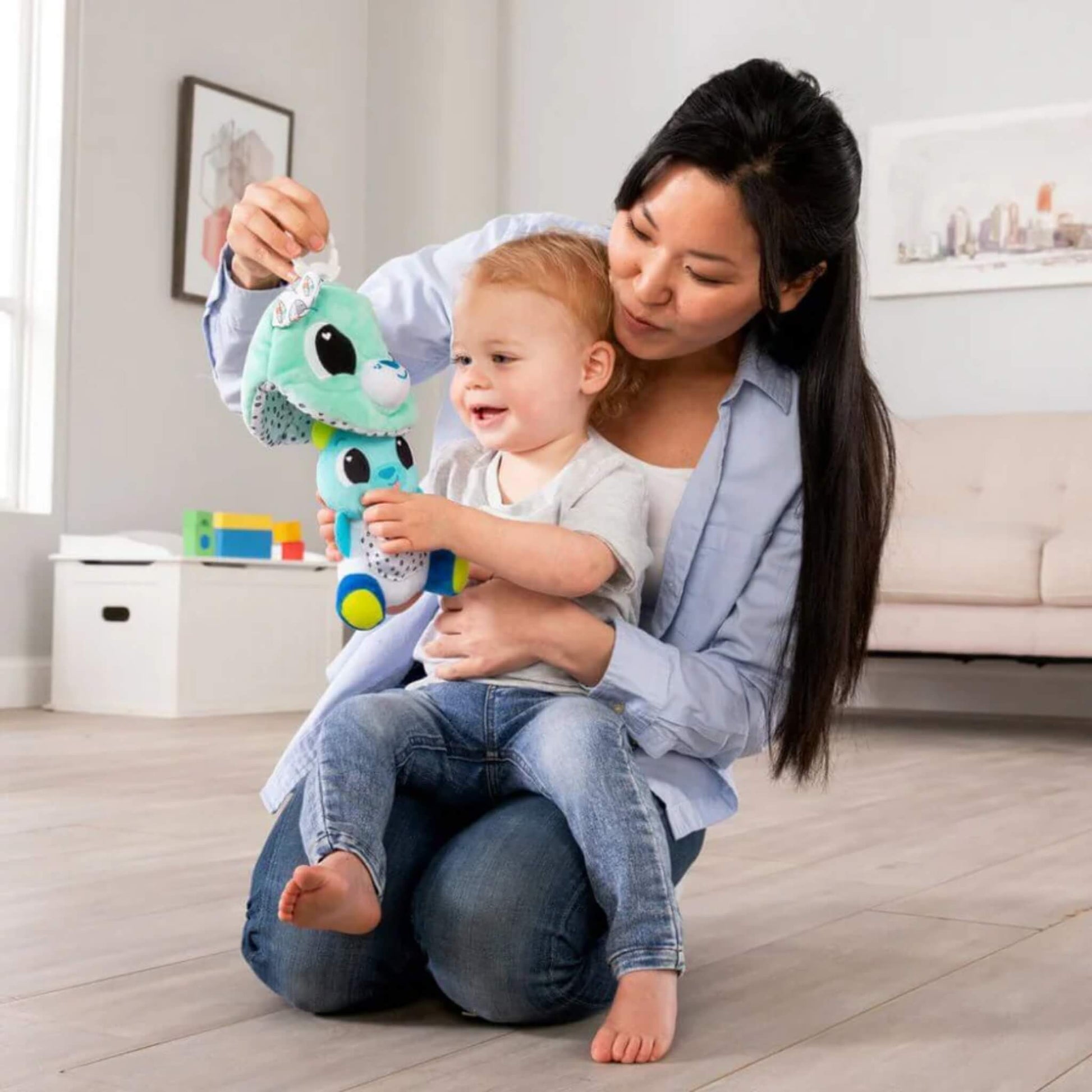 Mother sitting on the floor holding a plush bear toy while her toddler explores its colours and textures during playtime.