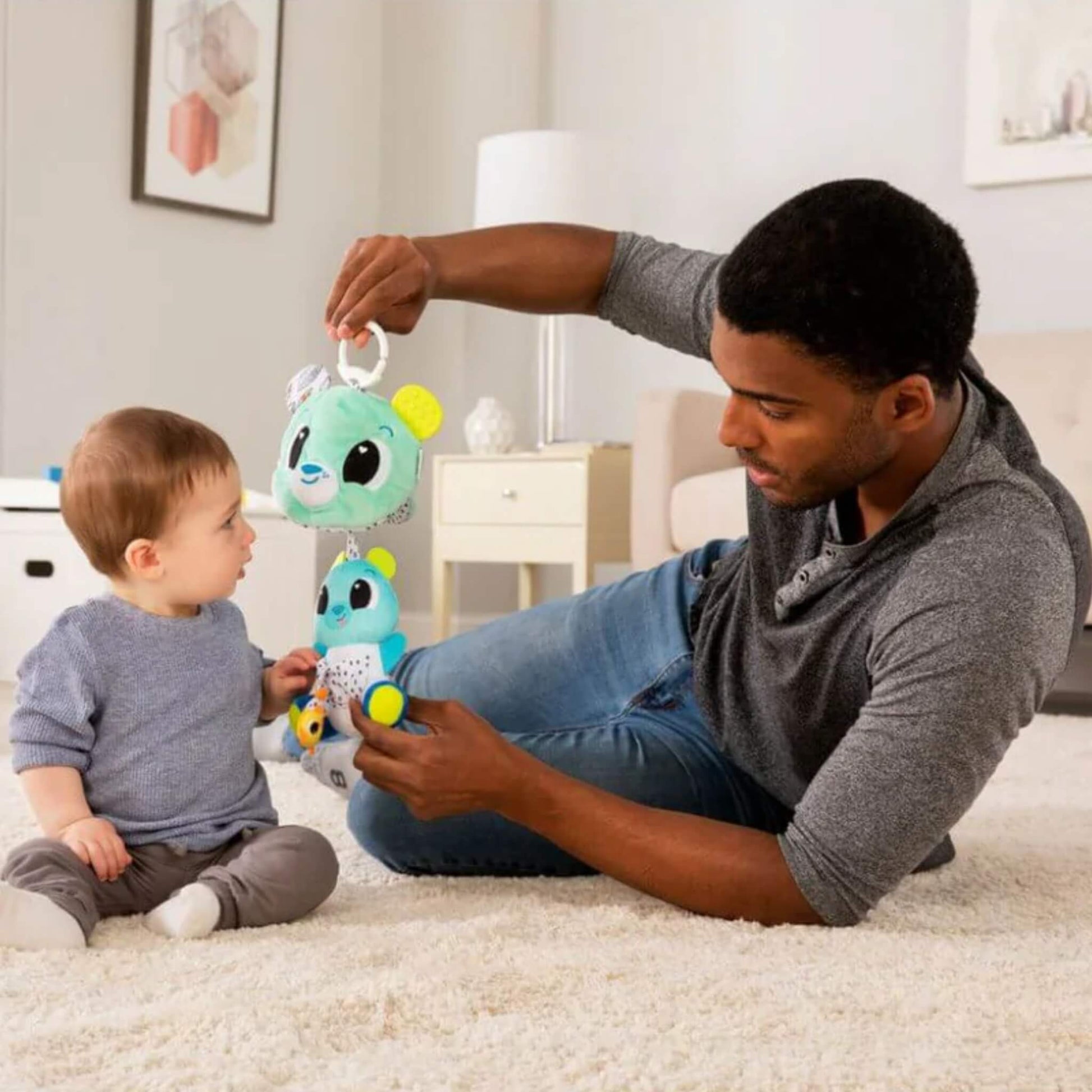 Father lying on a soft carpet interacting with his baby, holding a plush bear toy with layered bears for sensory discovery.