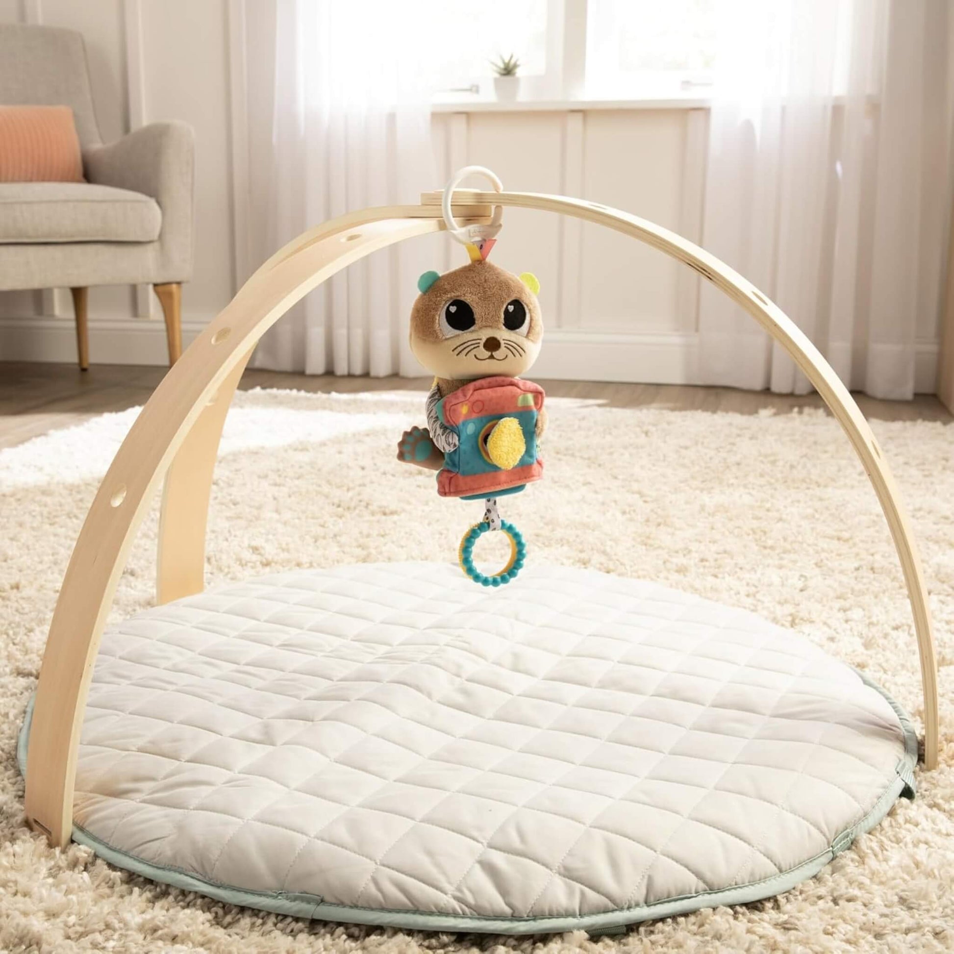 Fabric baby toy shaped like an otter hanging from a curved wooden play gym, positioned over a quilted round play mat.