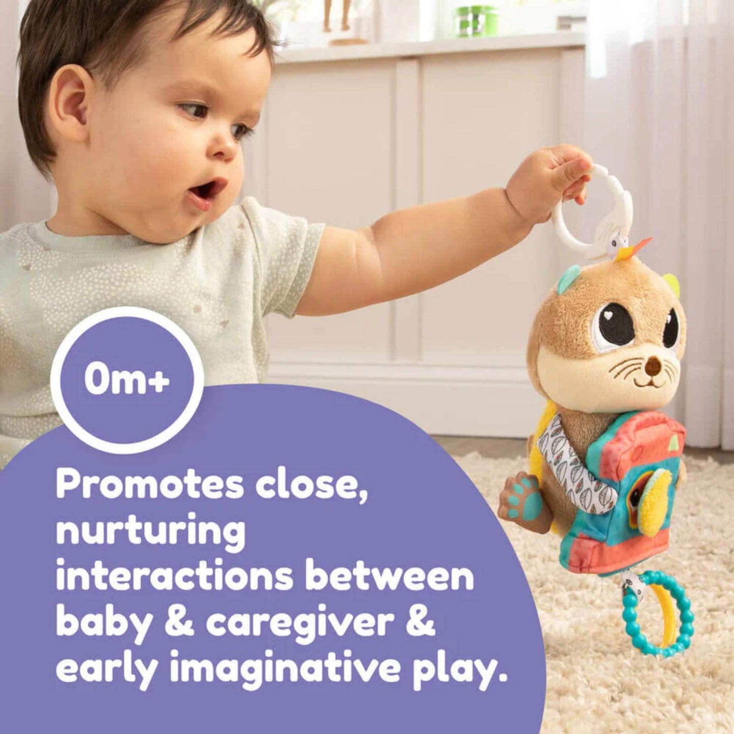 Baby lifting a plush otter toy by its clip while seated on a soft carpet, interacting with the toy during play.