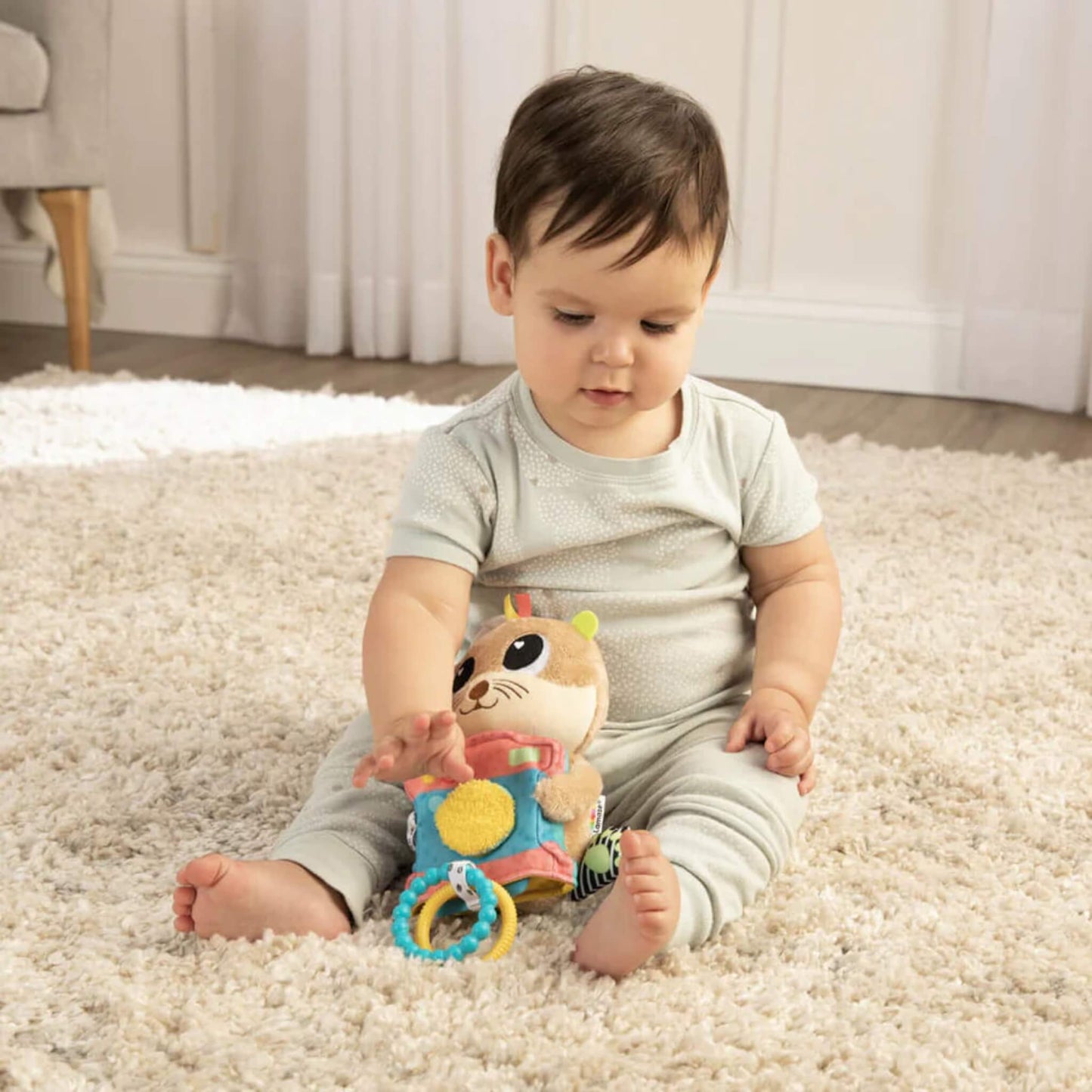 Baby seated on a neutral rug, gently touching a colourful plush toy designed for sensory exploration.