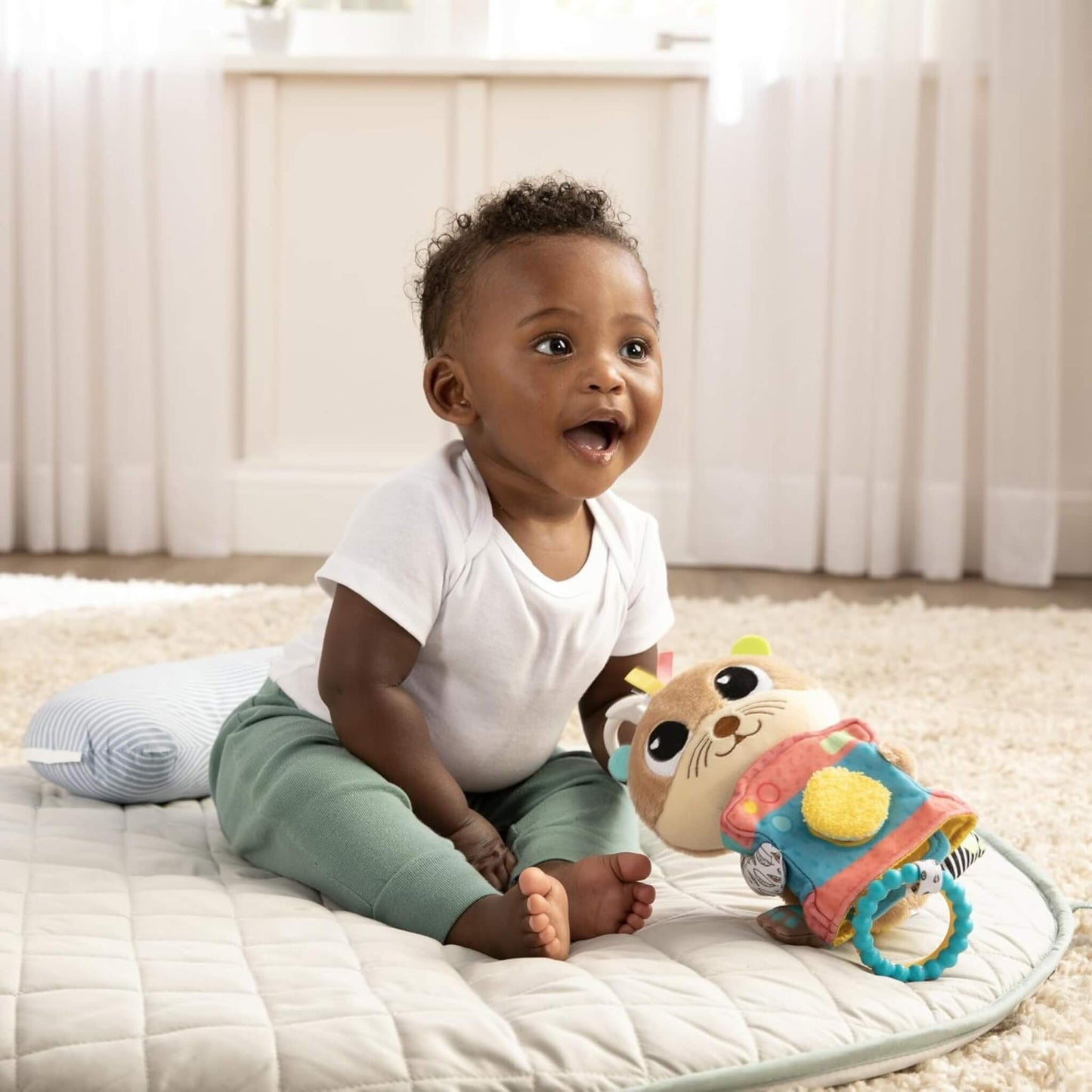 Smiling baby sitting on a padded quilted mat, holding a plush toy with a colourful camera design.