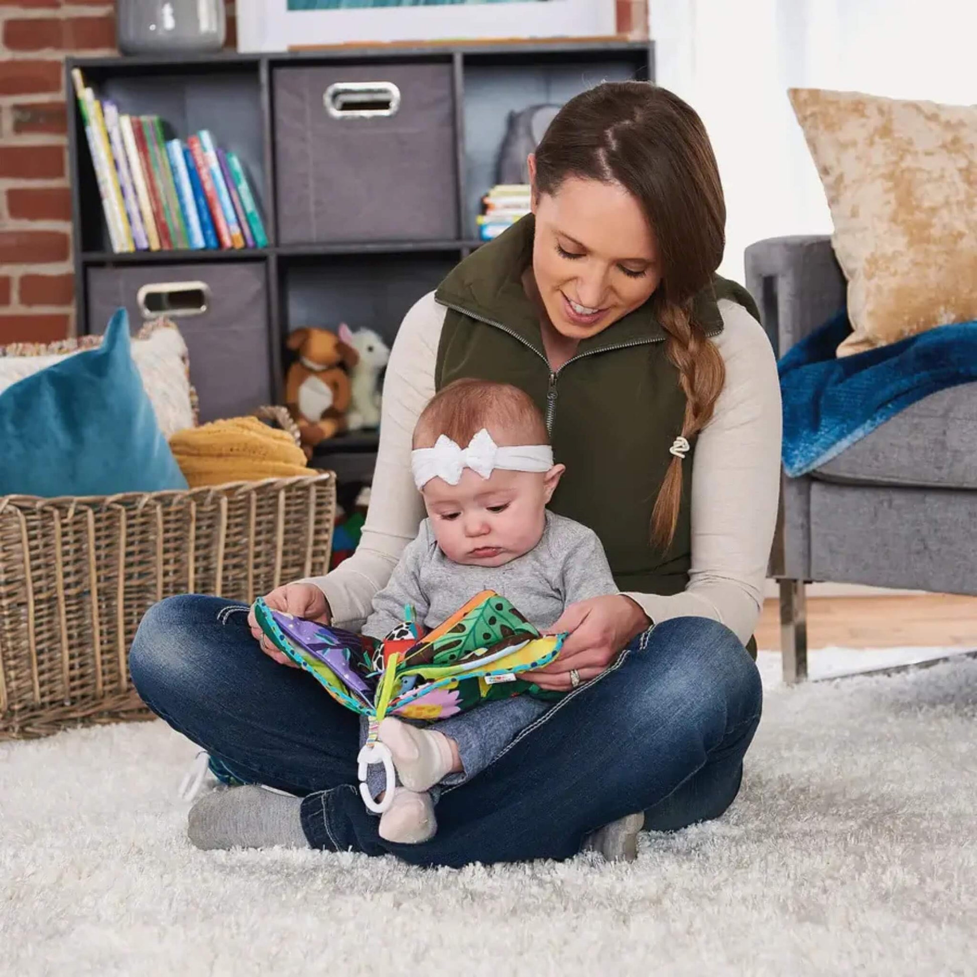 Mother sitting cross-legged on a soft rug, holding a baby in her lap as they look together at a colourful cloth baby book with bright pages and a small bug toy attached.
