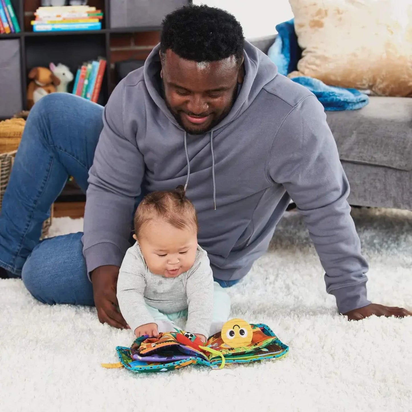 Father leaning beside a smiling baby on a white rug, both looking down at an open soft fabric baby book featuring colourful pages and a cheerful round character.