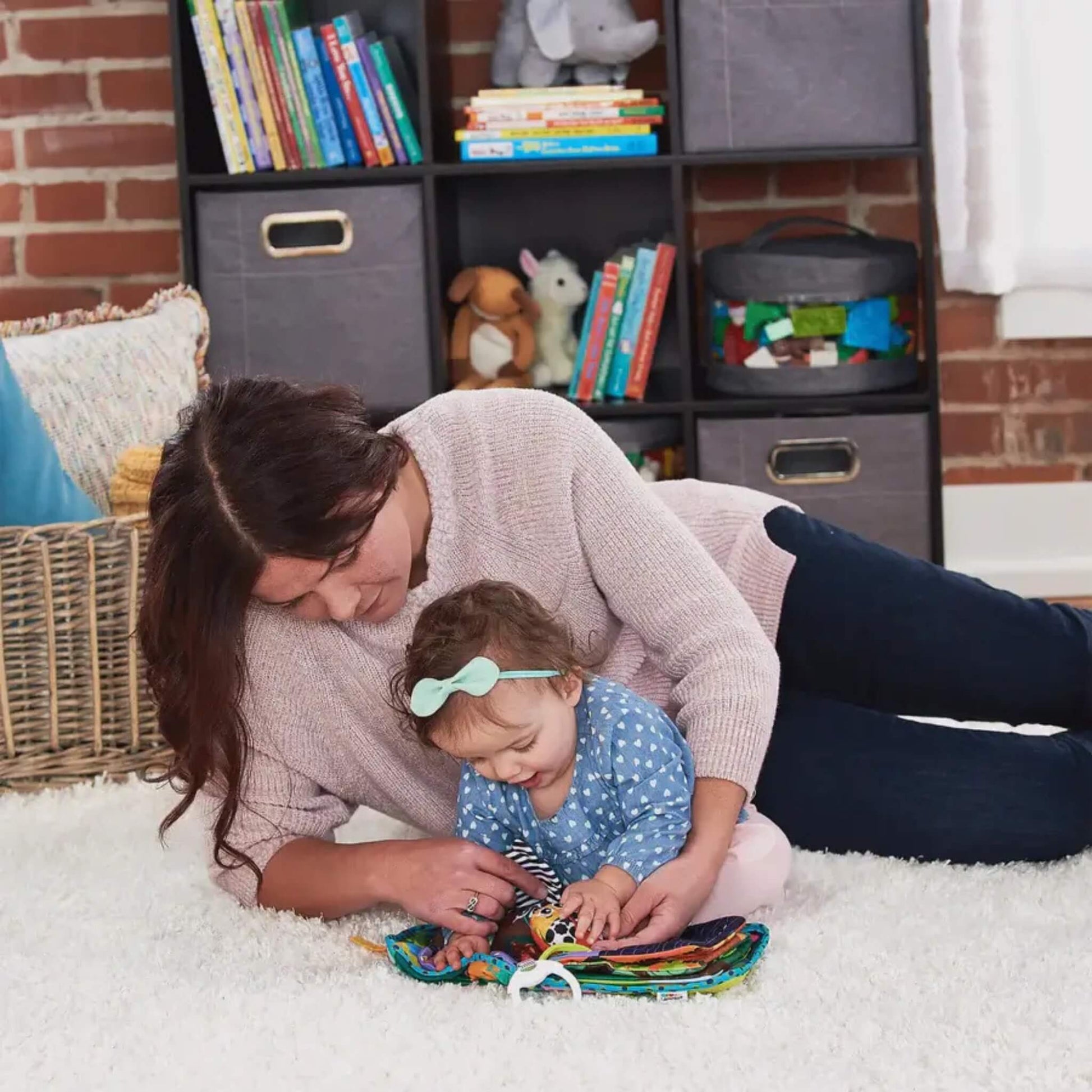 Mother lying on her side on a white rug with a toddler in her arms, helping the child turn the textured pages of a bright soft baby book with a small toy attached.