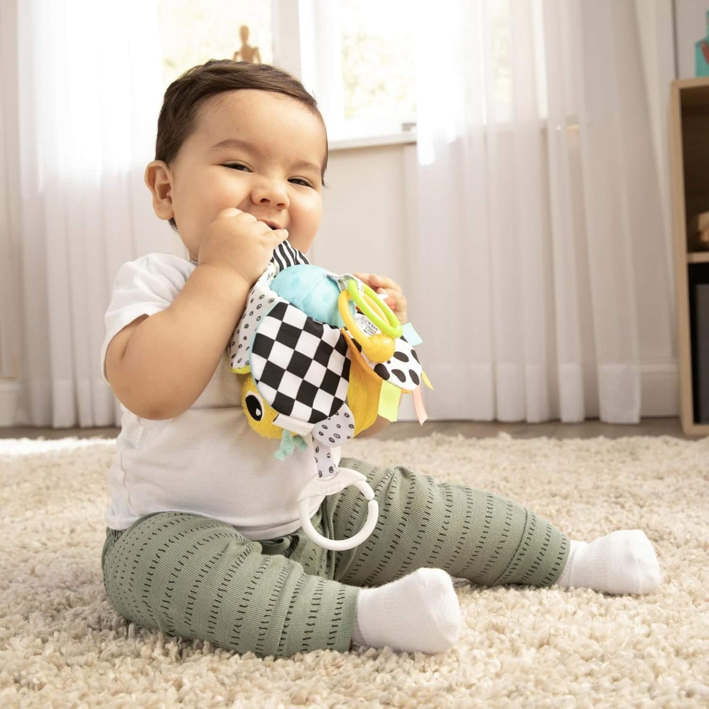 Baby sitting on a rug smiling while holding the bee toy and exploring it with their hands.