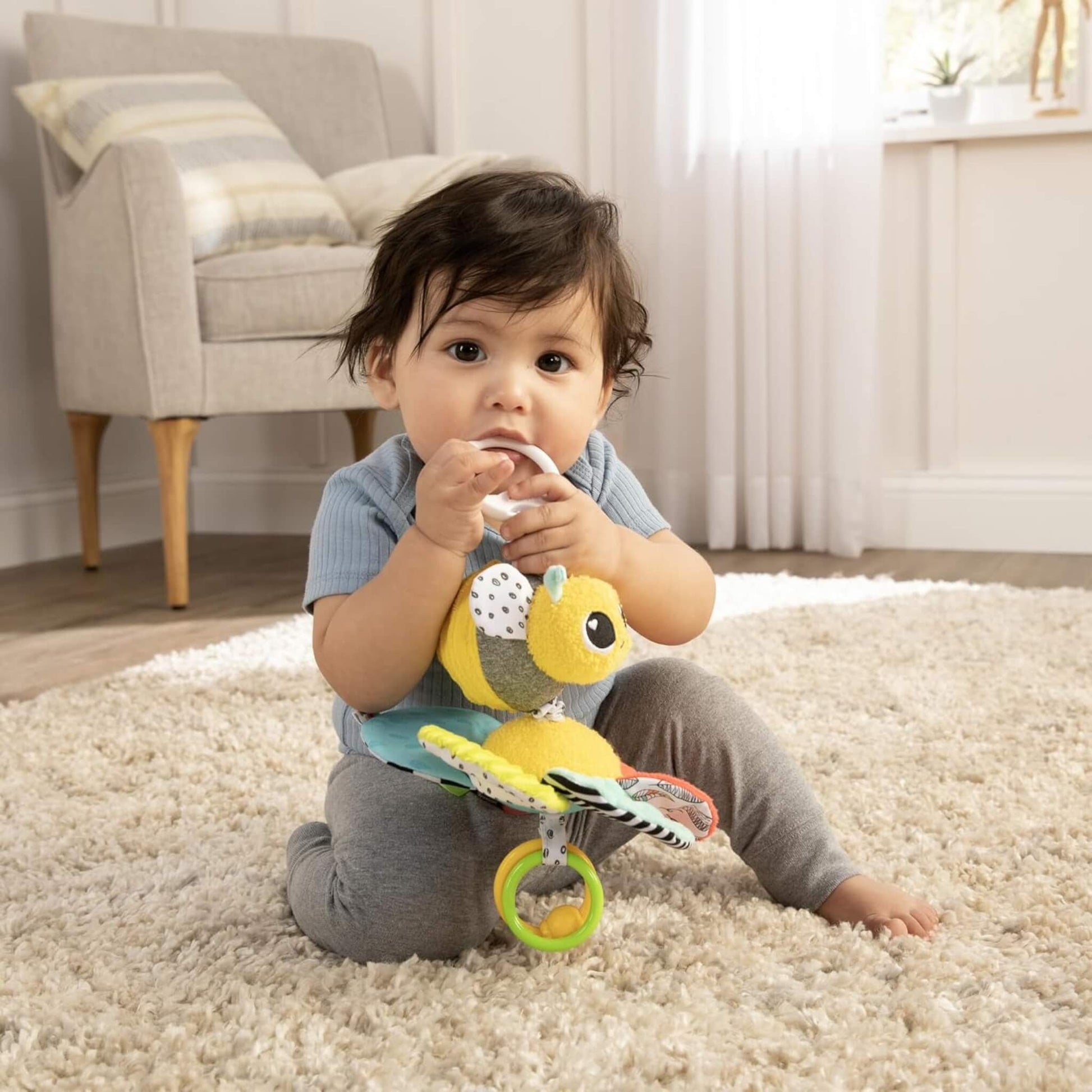 Baby seated on a carpet chewing the toy’s clip and exploring its soft petals.
