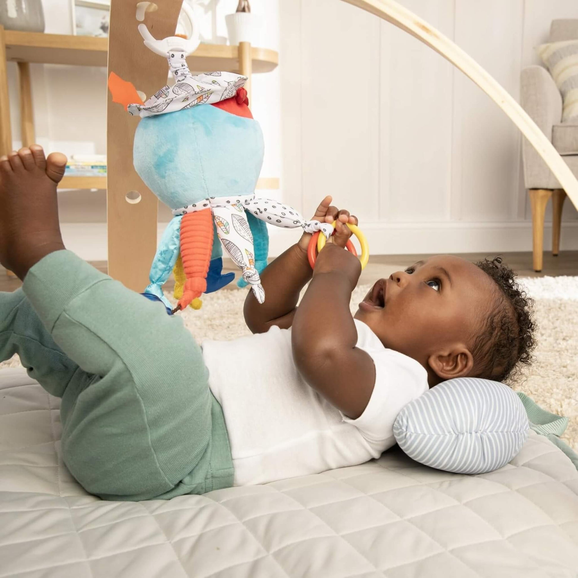 A baby lying on a play mat reaching up to grasp a hanging activity toy with fabric legs and clacking rings.