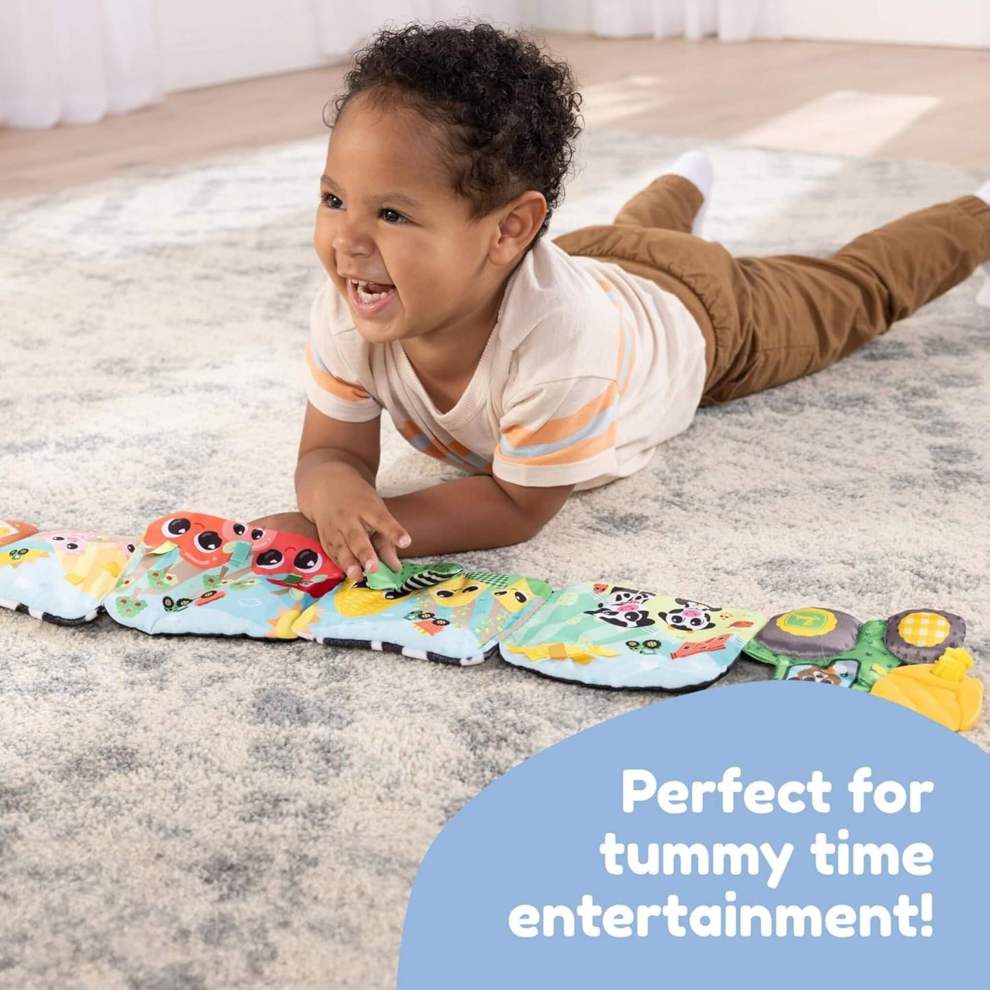 Baby lying on a soft rug during tummy time, exploring a fold-out fabric book decorated with animals and farm scenes.
