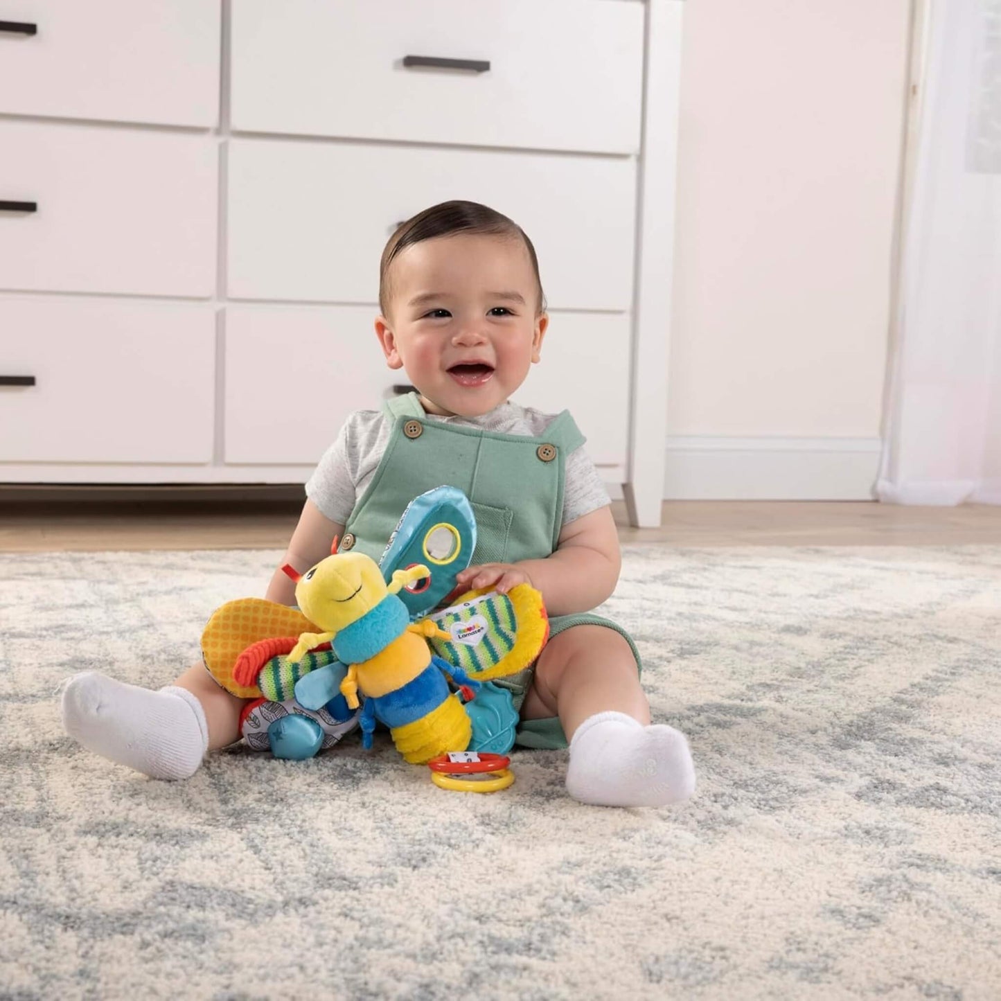 Baby sitting on a soft rug holding a plush firefly toy with large textured wings and bright patterns.