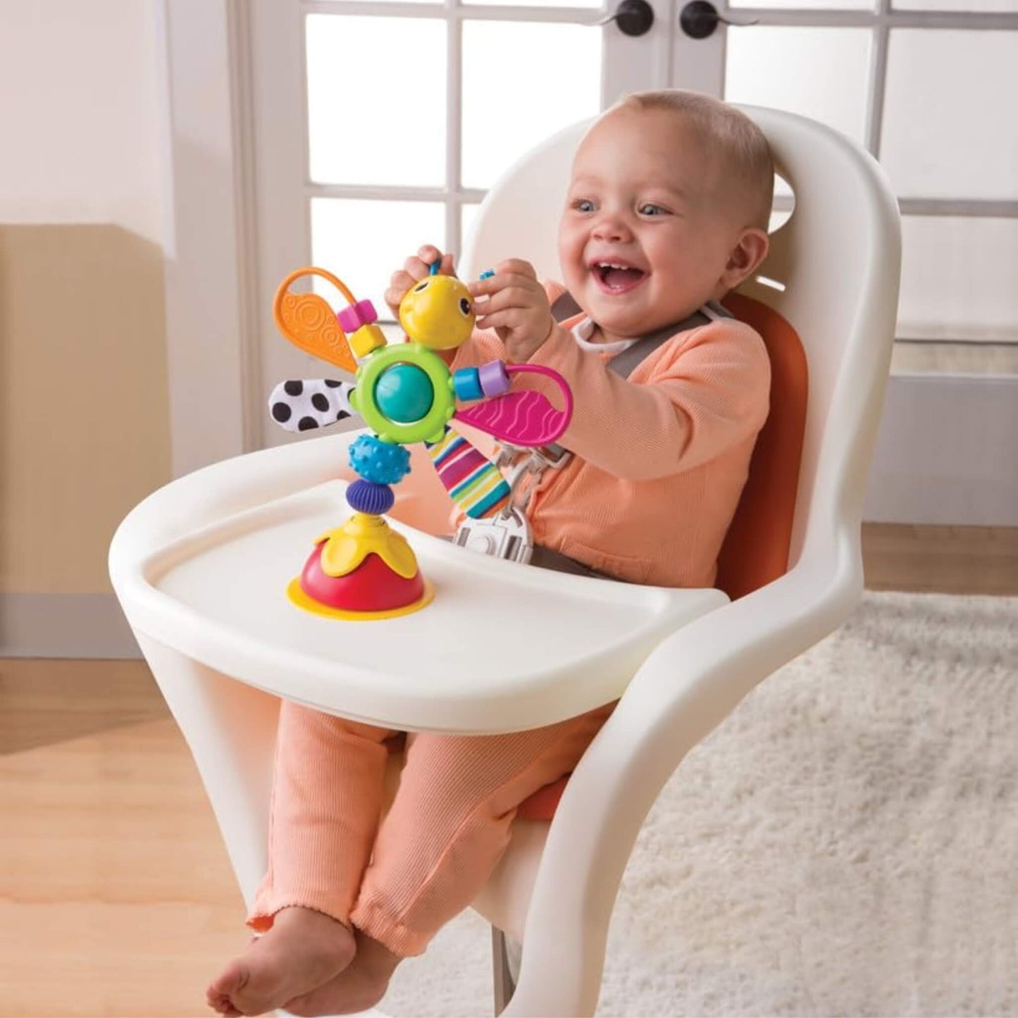 Baby sitting in a highchair, reaching out and smiling while spinning the colourful wings of a sensory toy on the tray.