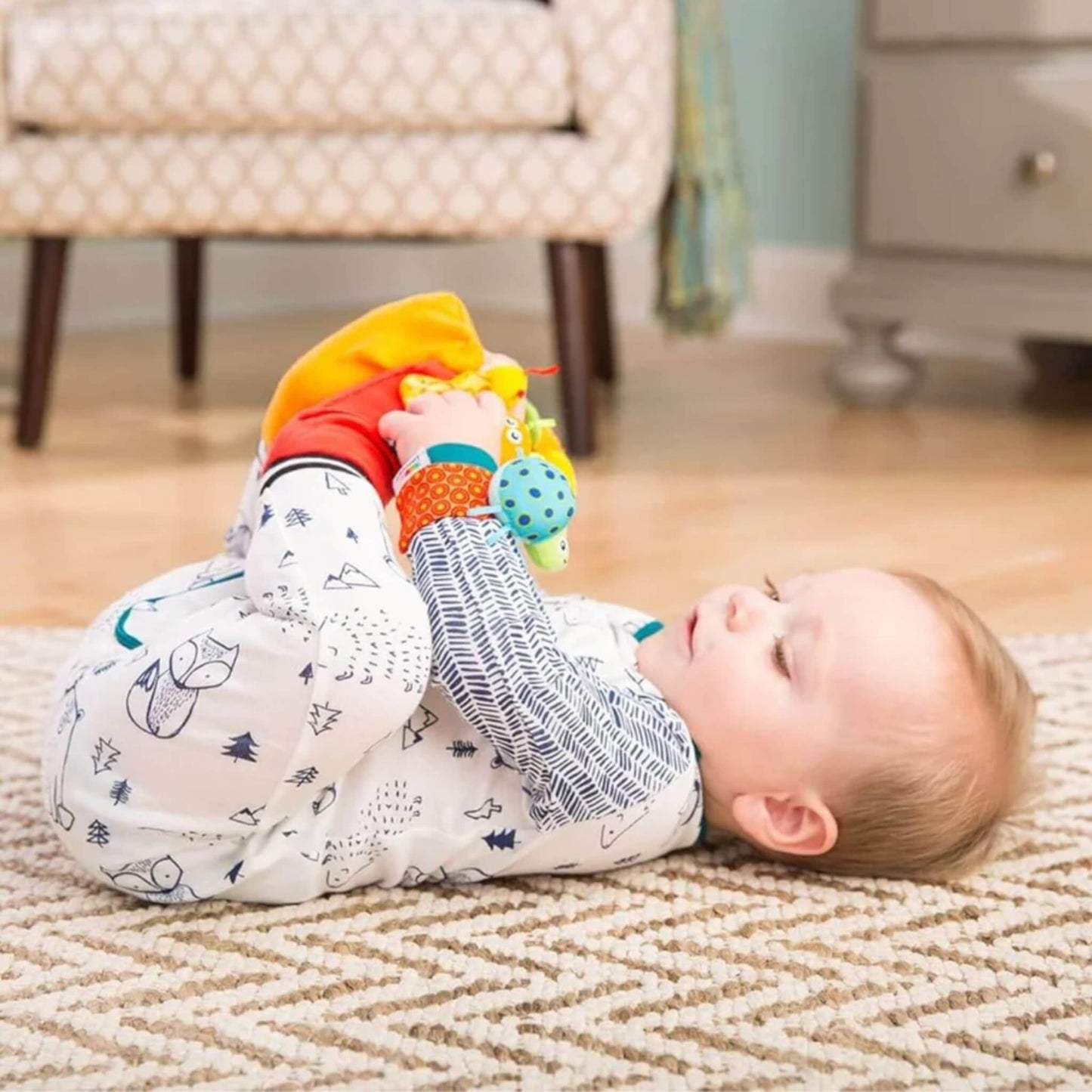 Baby lying on a patterned rug, wearing the footfinder socks and wrist rattles while lifting their feet to look at the attached bugs.