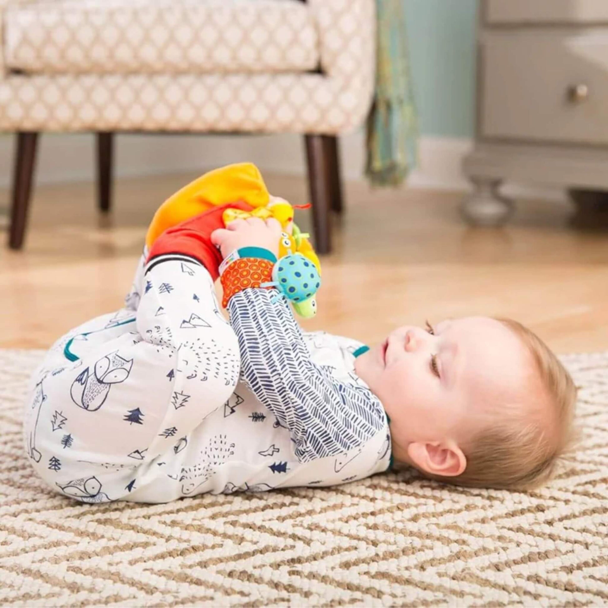 Baby lying on a patterned rug, wearing the footfinder socks and wrist rattles while lifting their feet to look at the attached bugs.