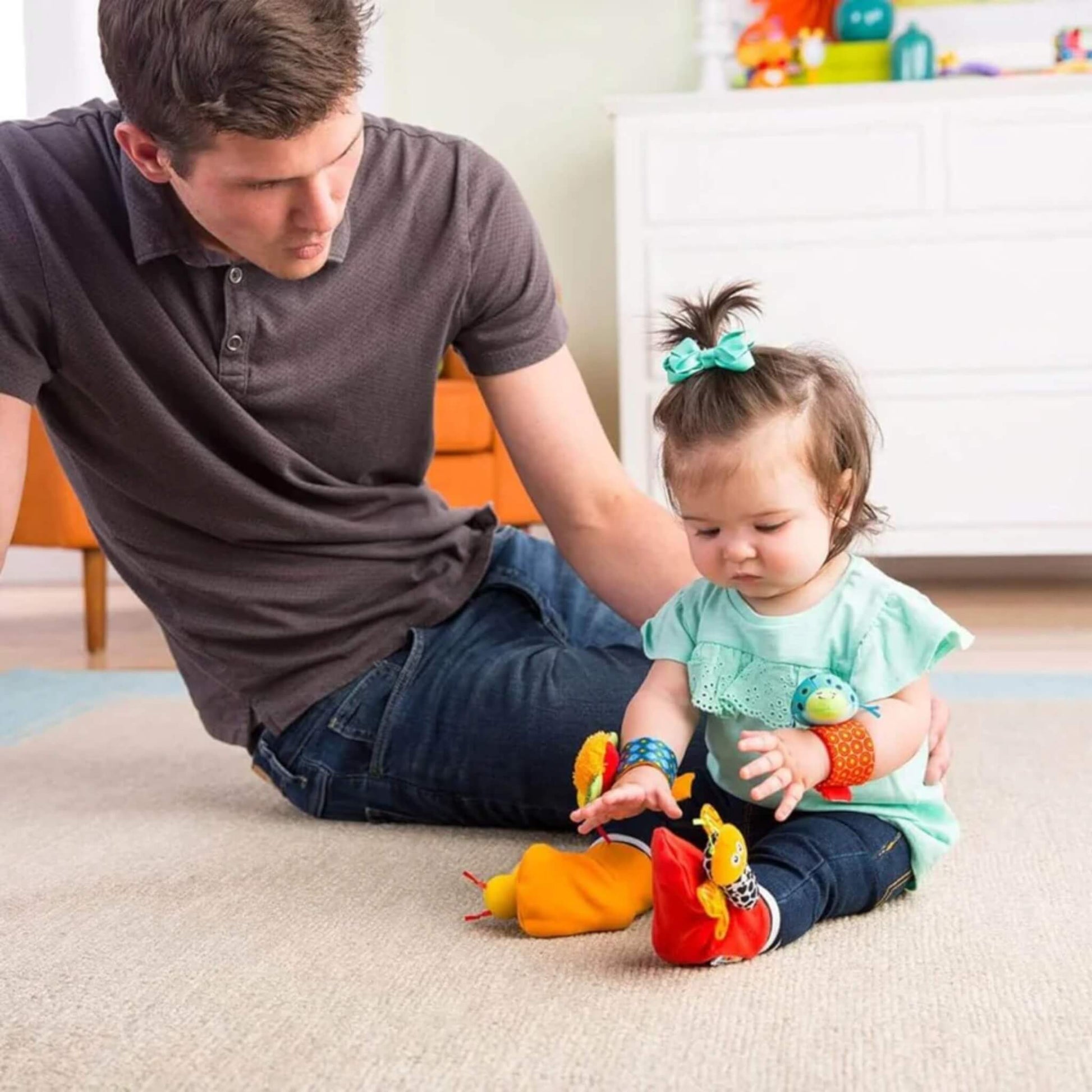 Parent sitting beside a baby who is wearing the wrist rattles and footfinders, both engaging in playtime on the floor together.