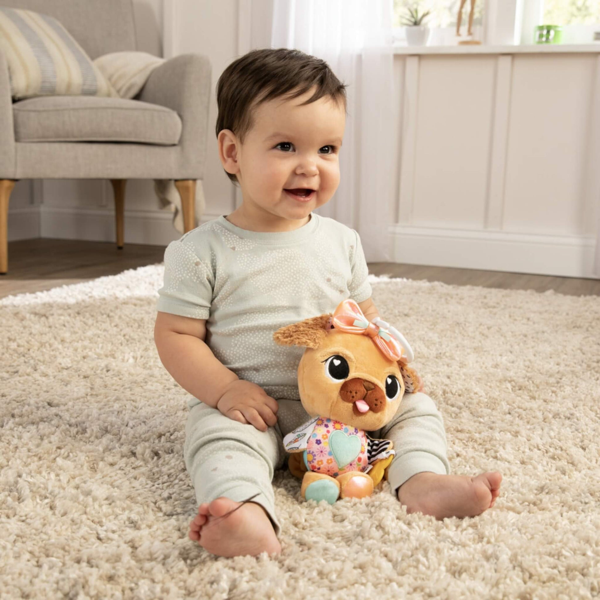 Baby sitting on a cream rug smiling while holding a plush pug toy with a bright bow and colourful fabric details.