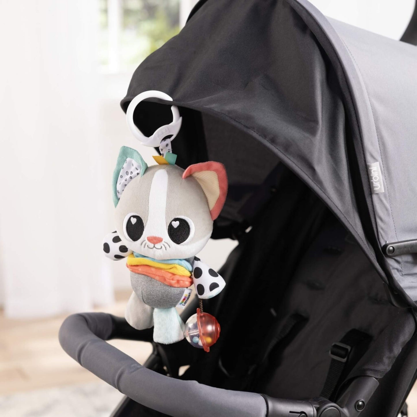 Plush sensory toy clipped securely to a stroller canopy handle, hanging within easy reach for a baby to play with.