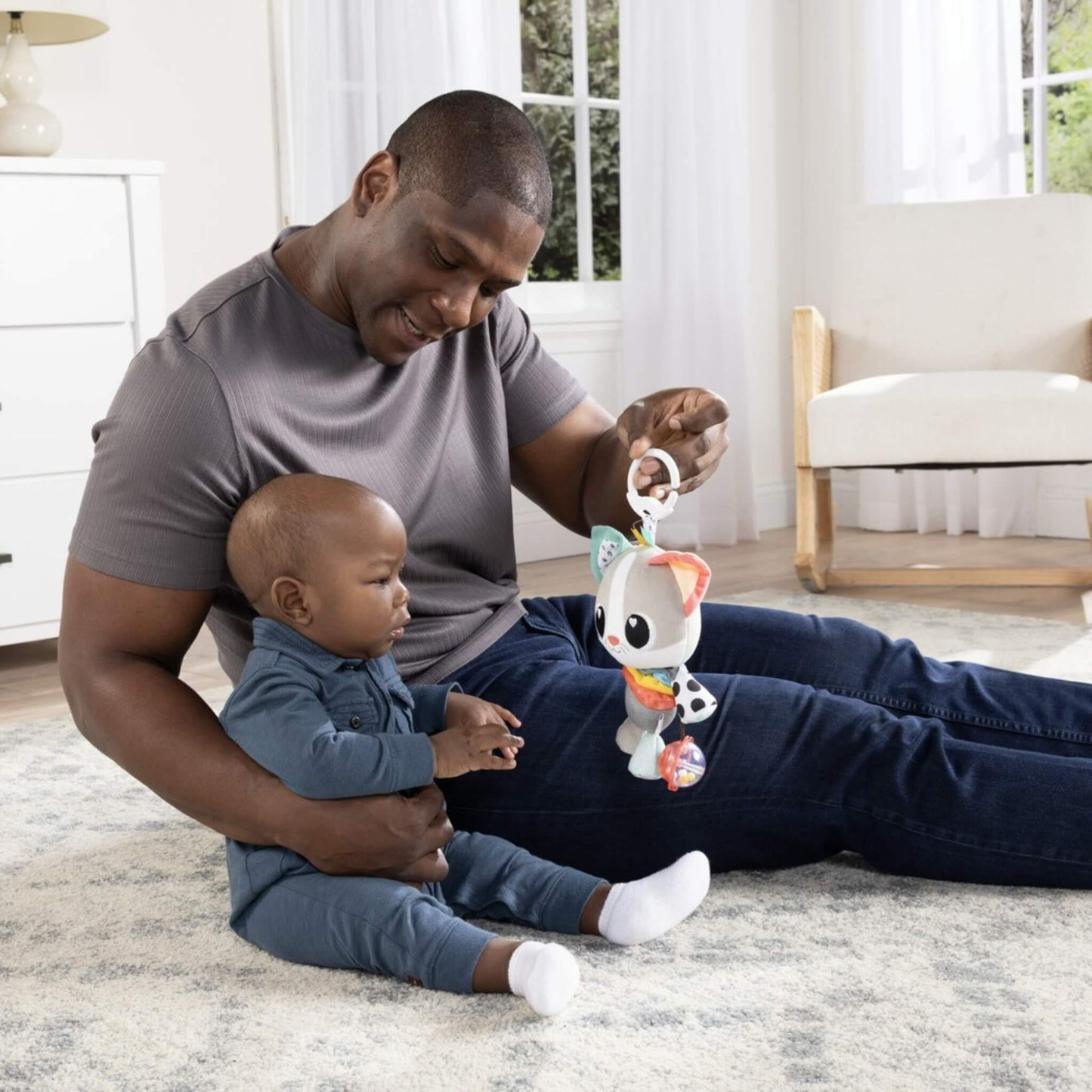 Father sitting on a soft rug holding a sensory kitten toy in front of his baby, encouraging touch and interaction.