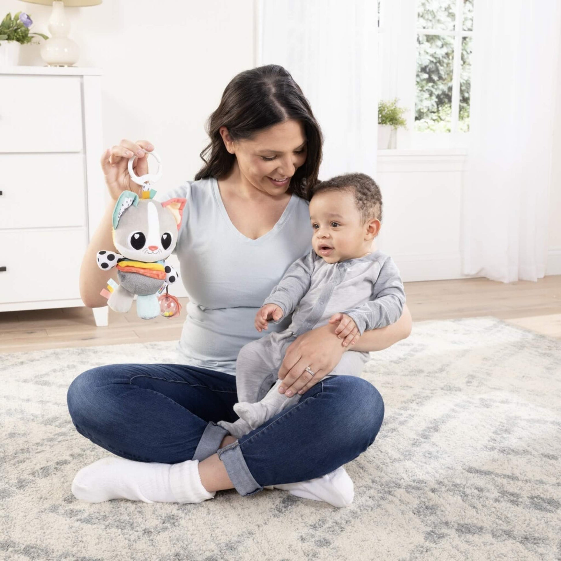 Mother seated on the floor with her baby, holding a plush activity toy with bold patterns and bright colours to capture attention.