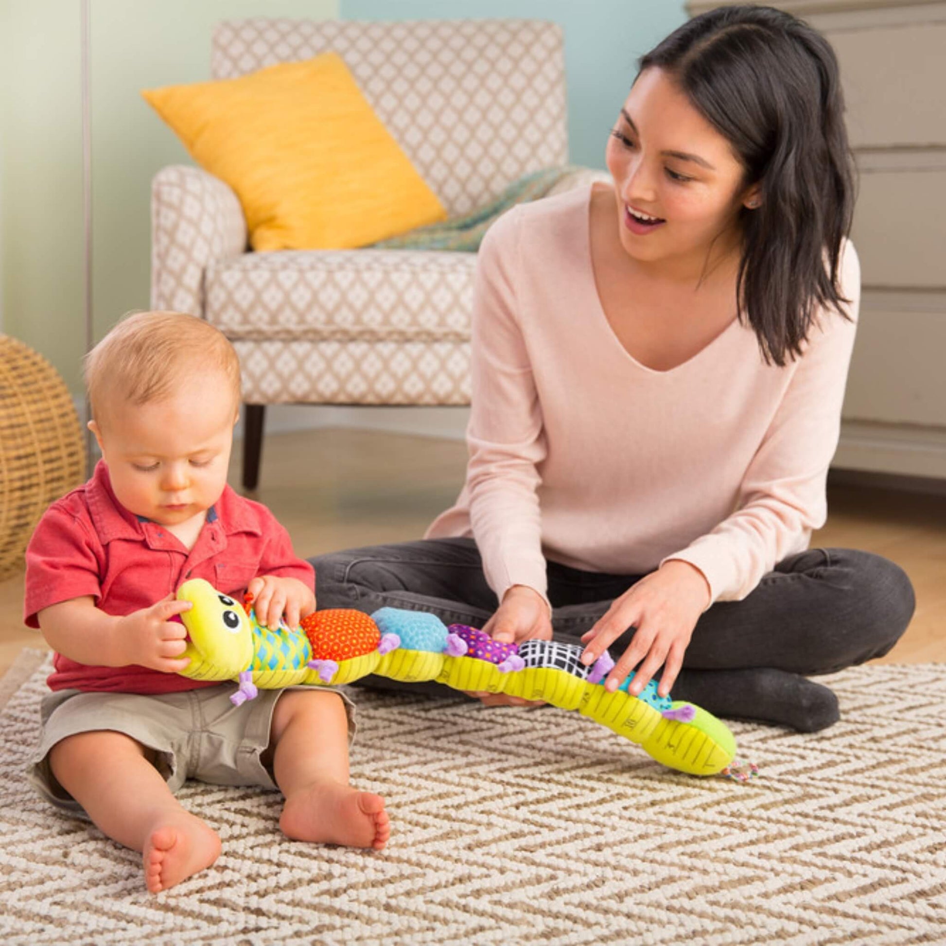 Mother sitting on the floor with her baby, both exploring a soft, brightly coloured inchworm toy laid out on a patterned rug.
