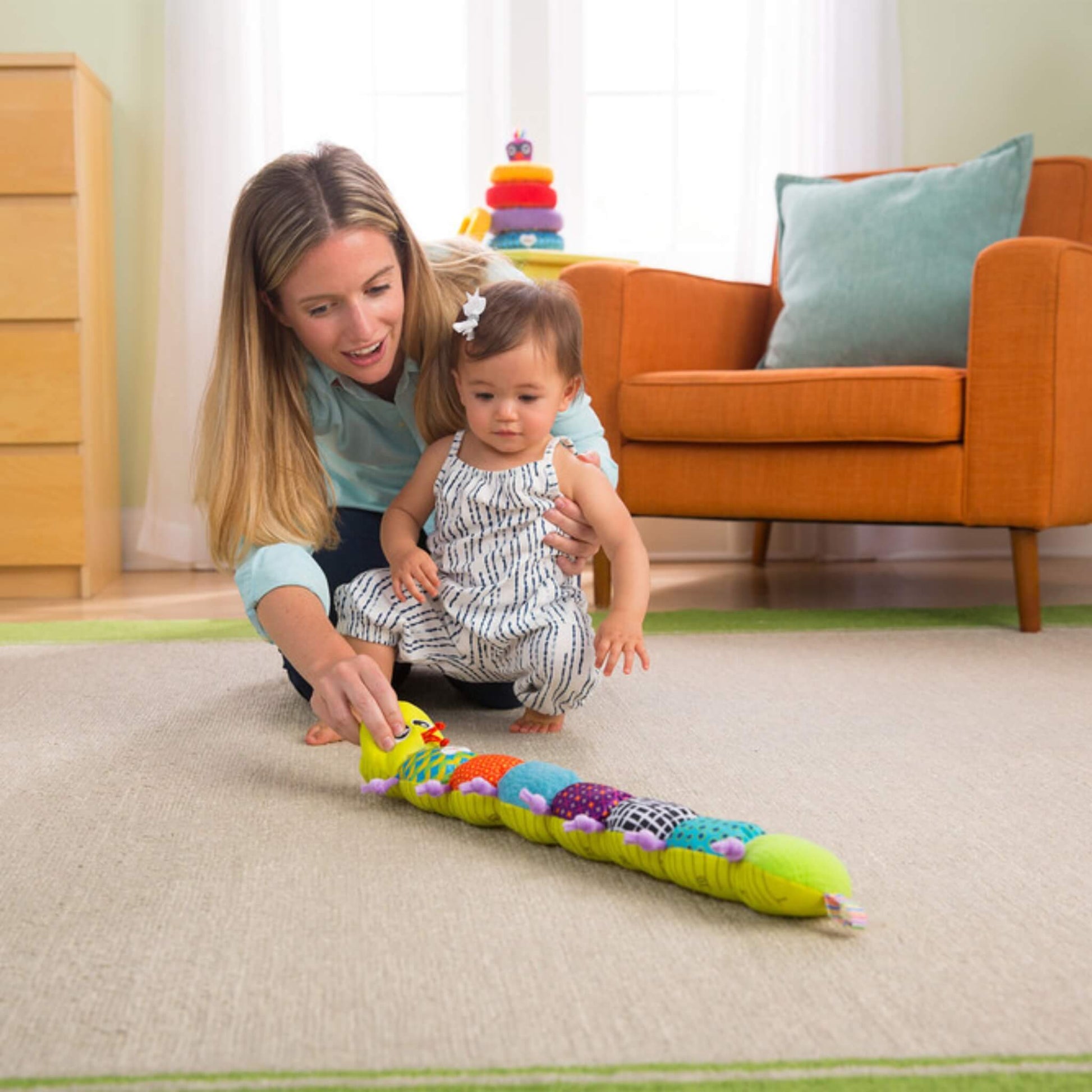 Mother and baby playing together on the floor, baby reaching forward to touch the inchworm toy’s face while mother encourages interaction.