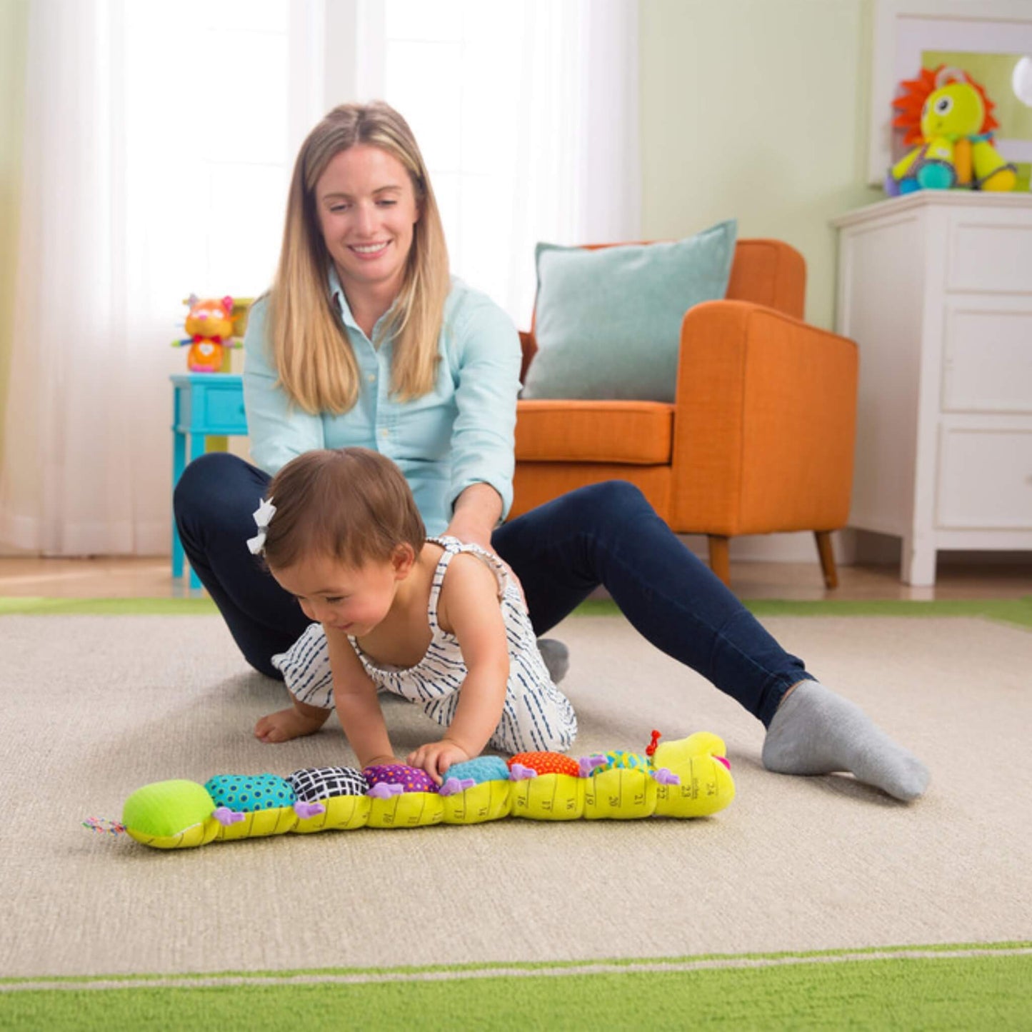 Mother seated on the floor behind her baby, who is leaning forward to press or explore the colourful musical inchworm toy.