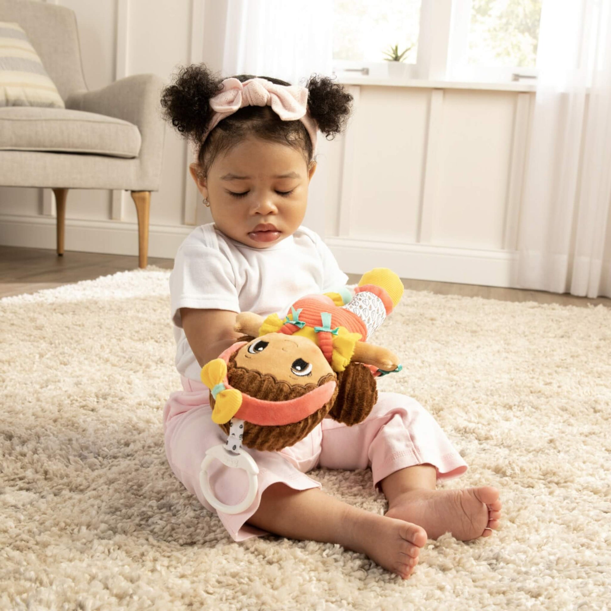 Baby sitting on a cream rug, holding a colourful soft doll with pigtails and bright clothing while exploring its textures and details.