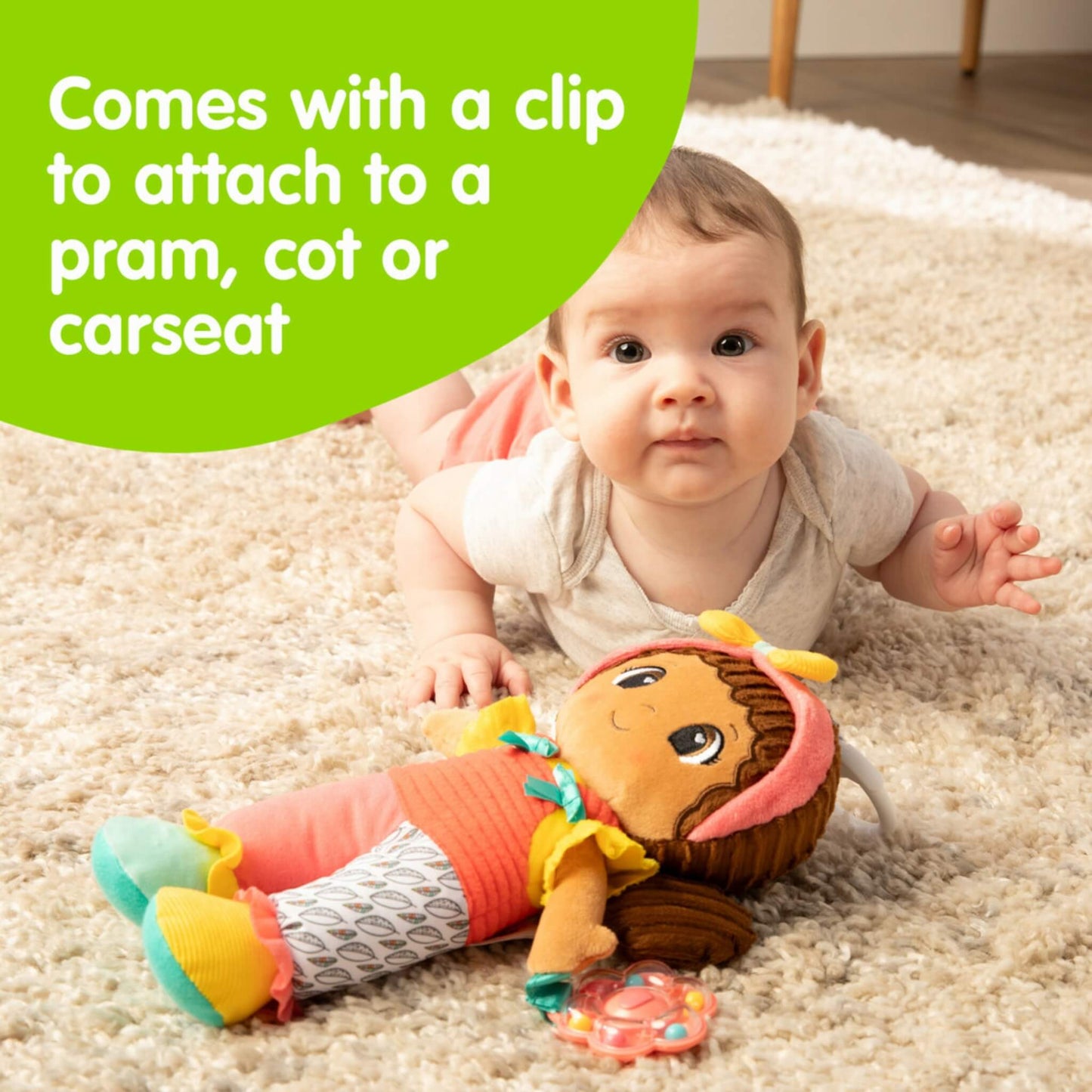Baby lying on the floor next to a bright Lamaze doll with text pointing out the clip feature that allows attachment to a pram, cot or car seat.